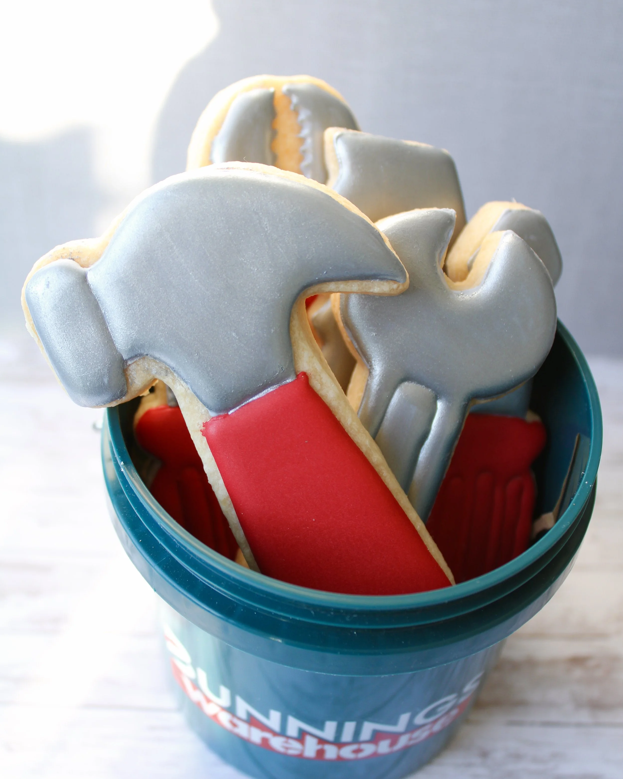 Close up of Father's Day tool cookies decorated in silver and red icing, presented in a dark green Bunnings Warehouse mini bucket on a grey washed wooden background.