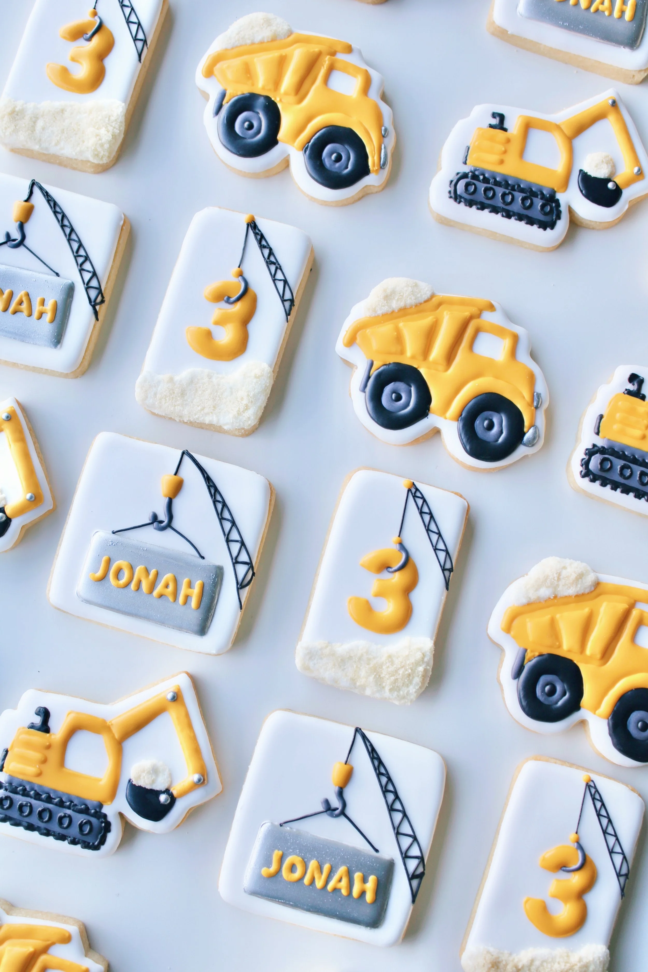 Construction Birthday Cookie Set featuring trucks, excavators and cranes in golden yellow, white, silver and black with personalised name and age cookies.