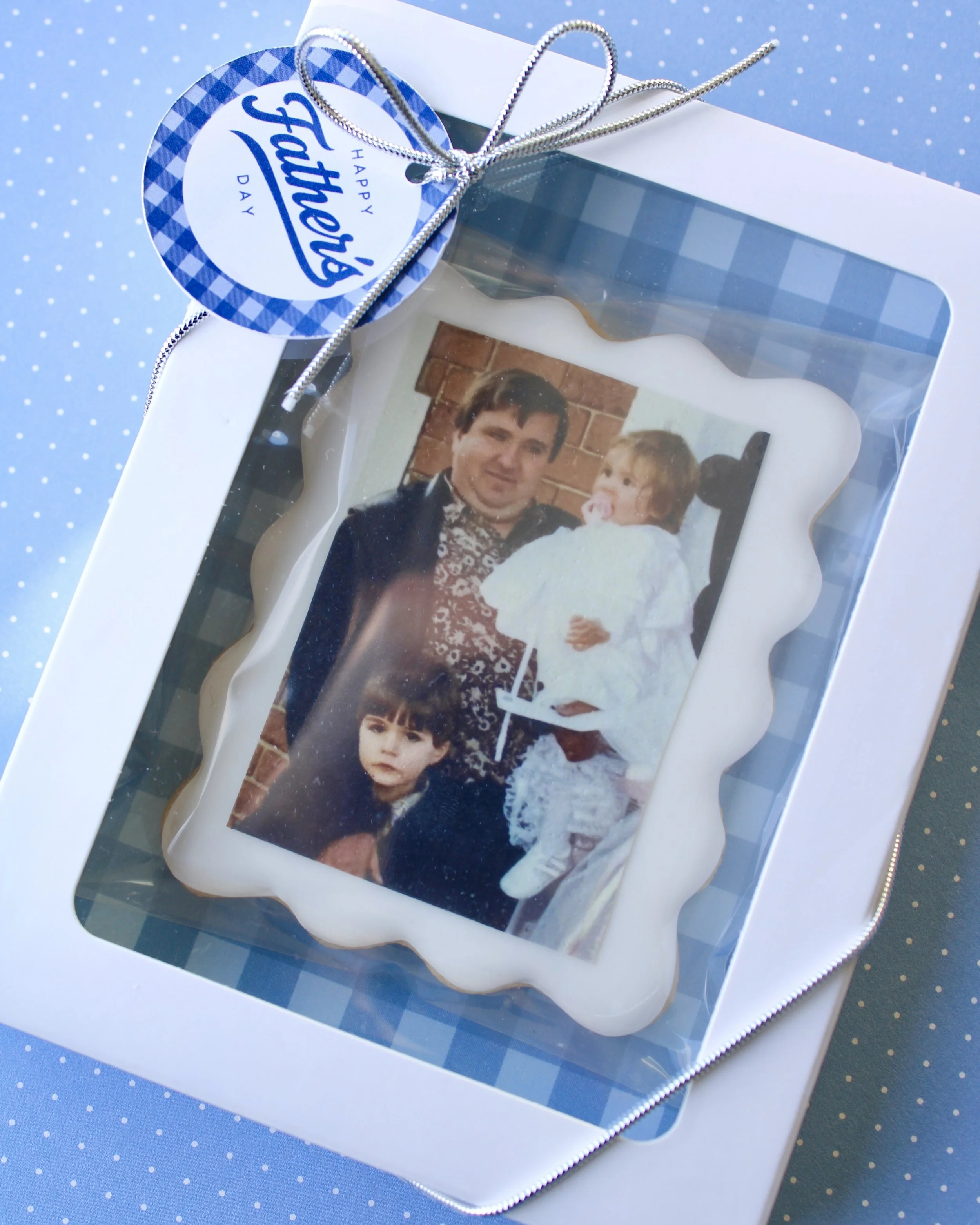 Personalised Father’s Day photo cookie gift box with white iced cookie, wavy border, printed photo, blue gingham backing, Father’s Day gift tag, and ribboned white cookie box on a blue background.
