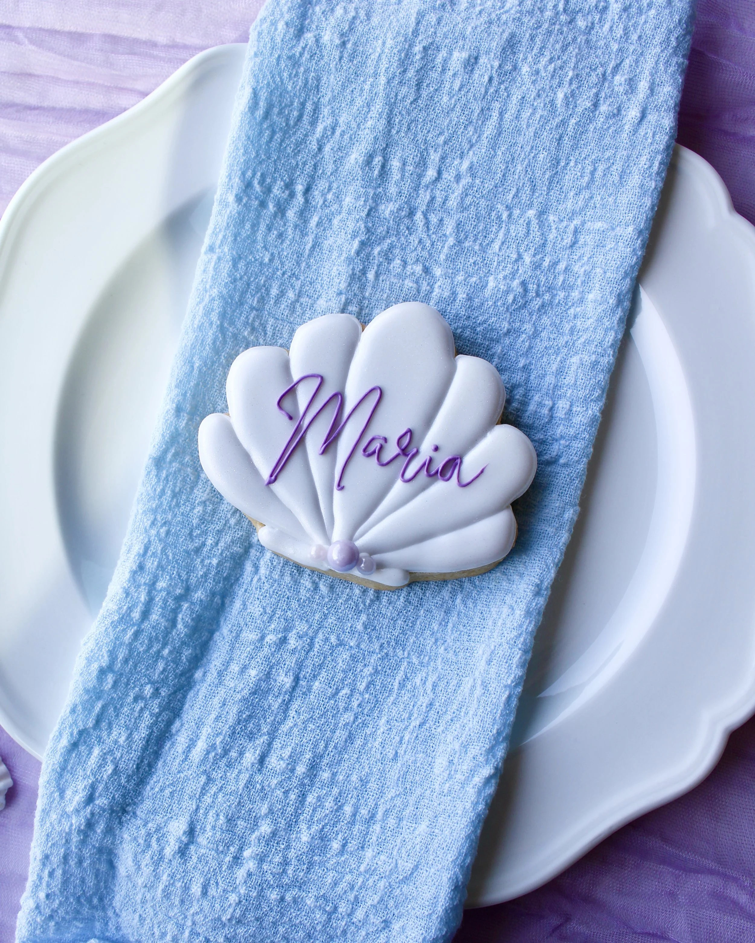 White seashell cookie with the name “Maria” written in purple cursive icing, placed on a white vintage plate over blue linen napkin and purple crinkled fabric with scattered seashells.