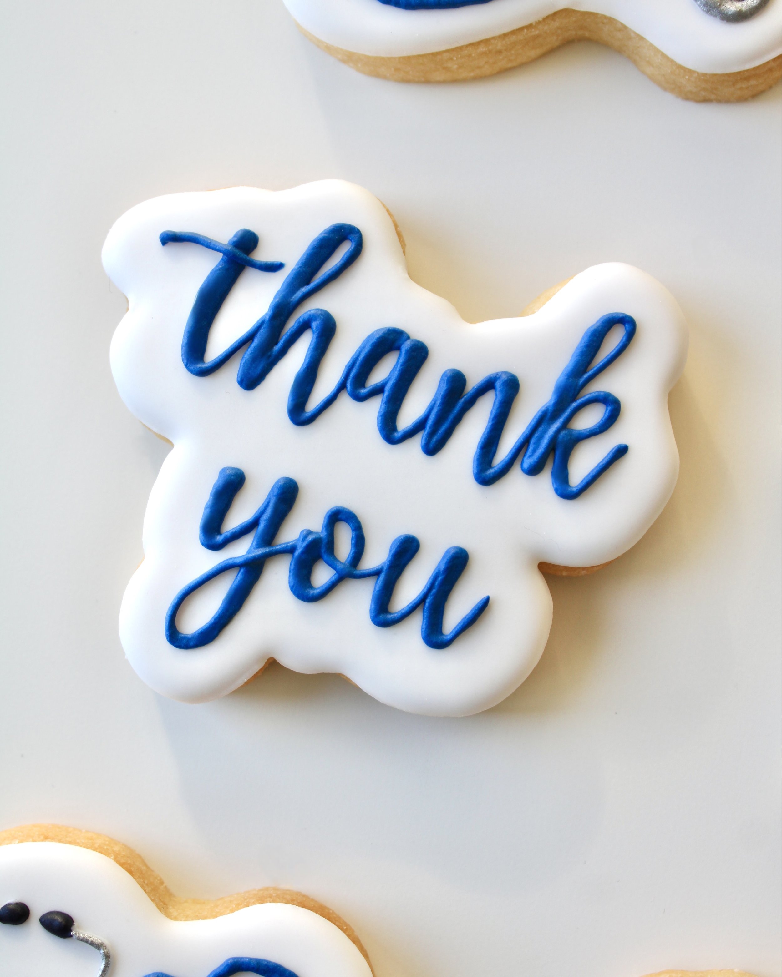 Close-up of a white iced cookie with the words “thank you” written in dark blue cursive icing.