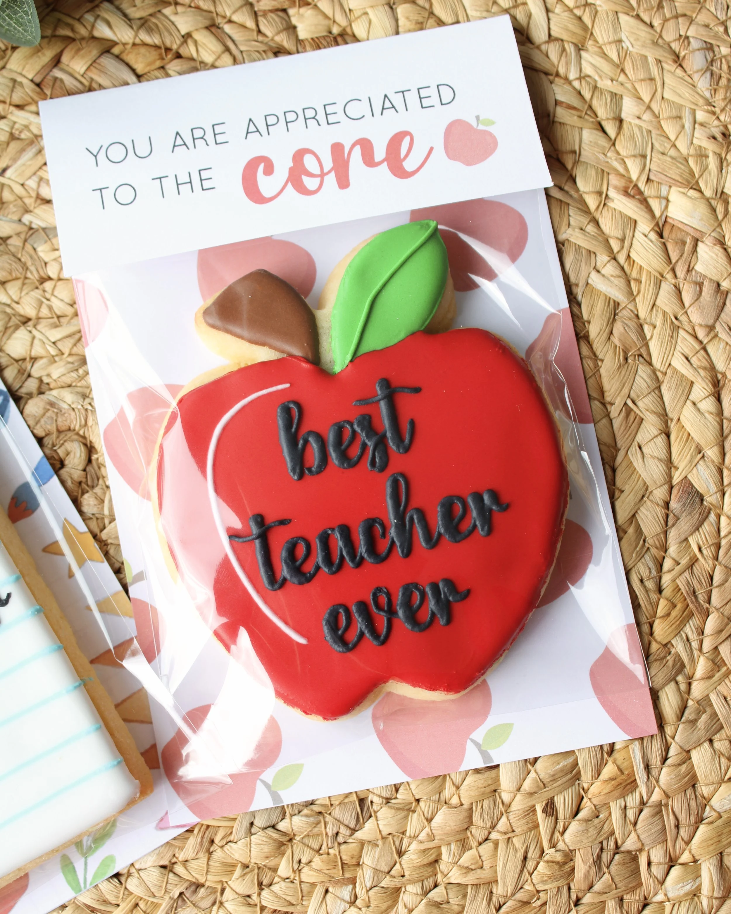 Close-up of a teacher appreciation cookie on a hyacinth placemat with eucalyptus. Red apple cookie with green leaf and “best teacher ever” icing, topped with “You are appreciated to the core.”