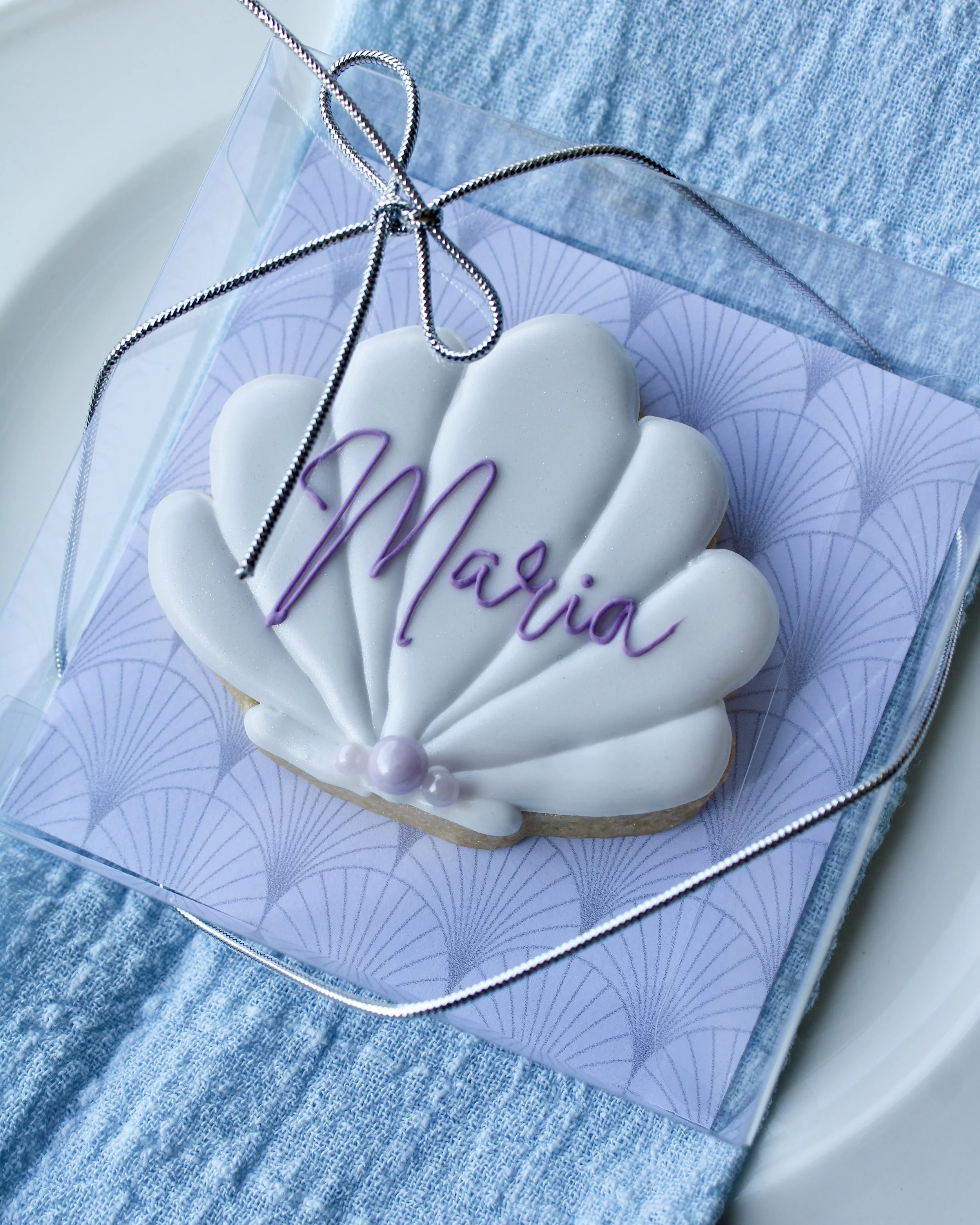 White seashell cookie with “Maria” in purple cursive icing, in a clear box tied with silver cord bow, on a white vintage plate over blue linen and purple crinkled fabric with seashells.