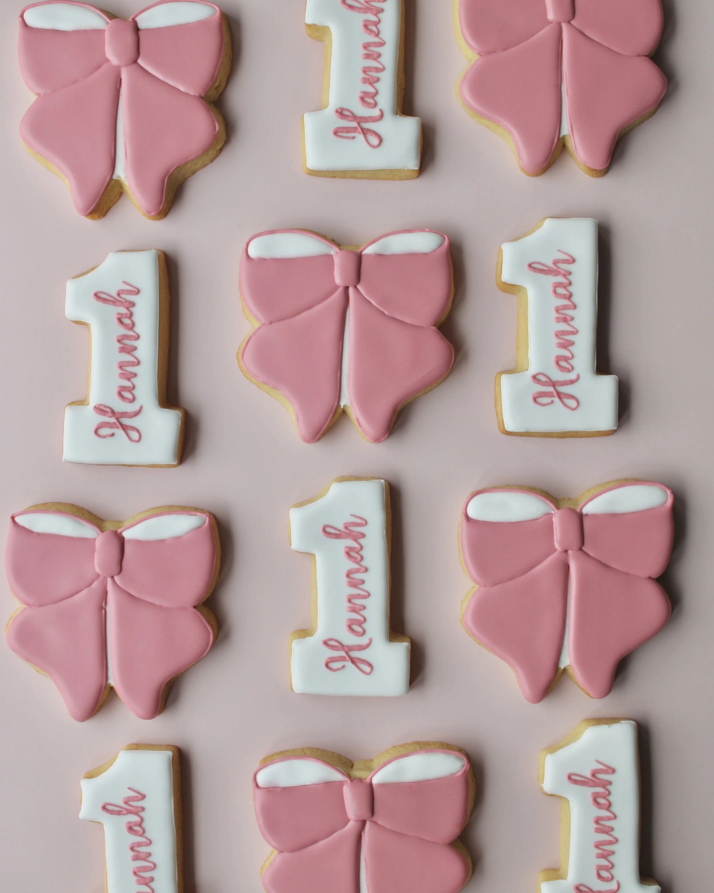 Pink and white cookie set featuring a white number 1 cookie with “Hannah” written in blush script and blush bow cookies for the remaining cookies.