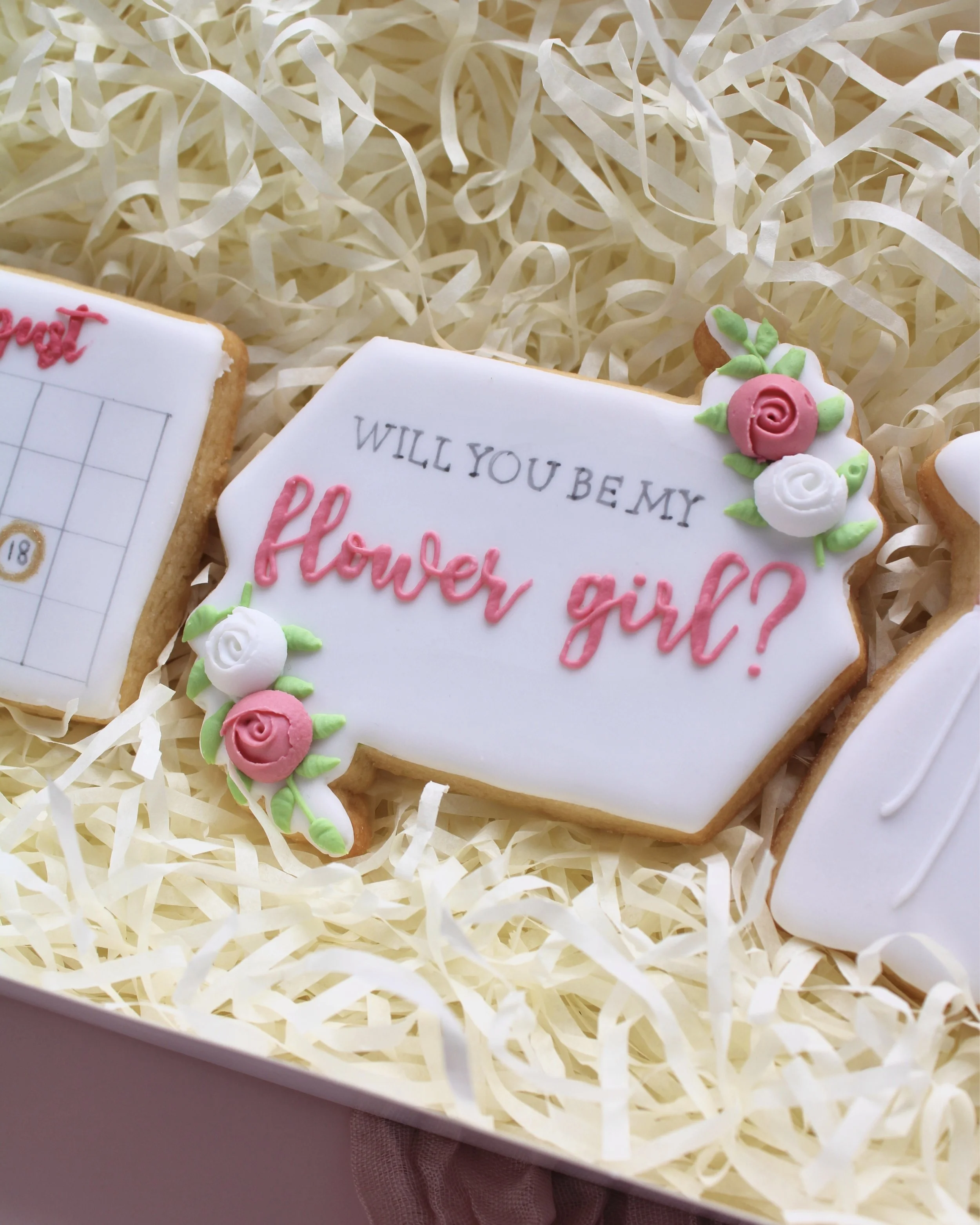 Flower girl cookie proposal box featuring three cookies, close-up of a white elongated hexagon cookie with the words “will you be my flower girl” and pink and white rose details.