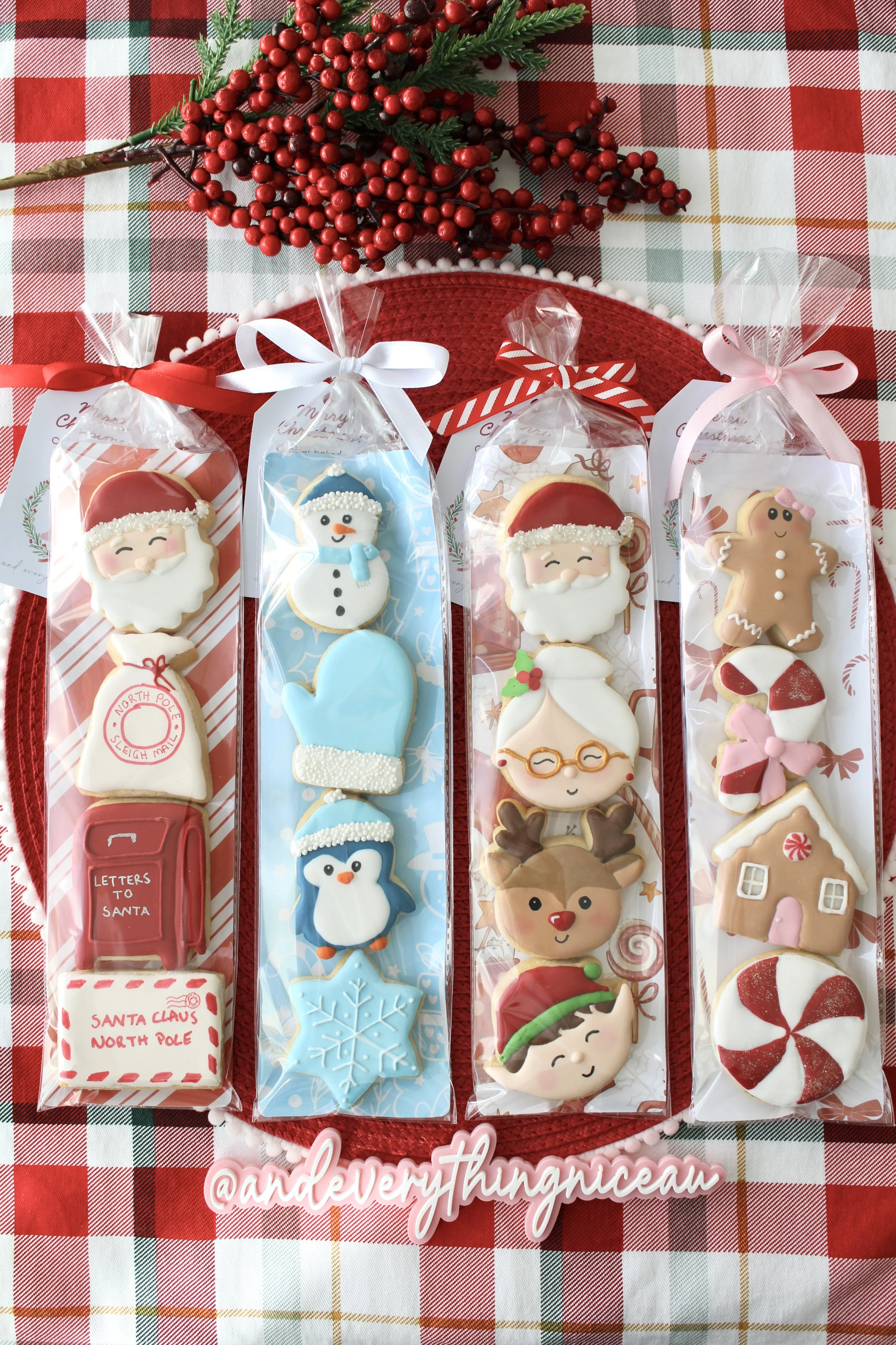 Four Christmas cookie packs, each in a different holiday-themed sleeve, tied with ribbons, styled on red and green tartan fabric