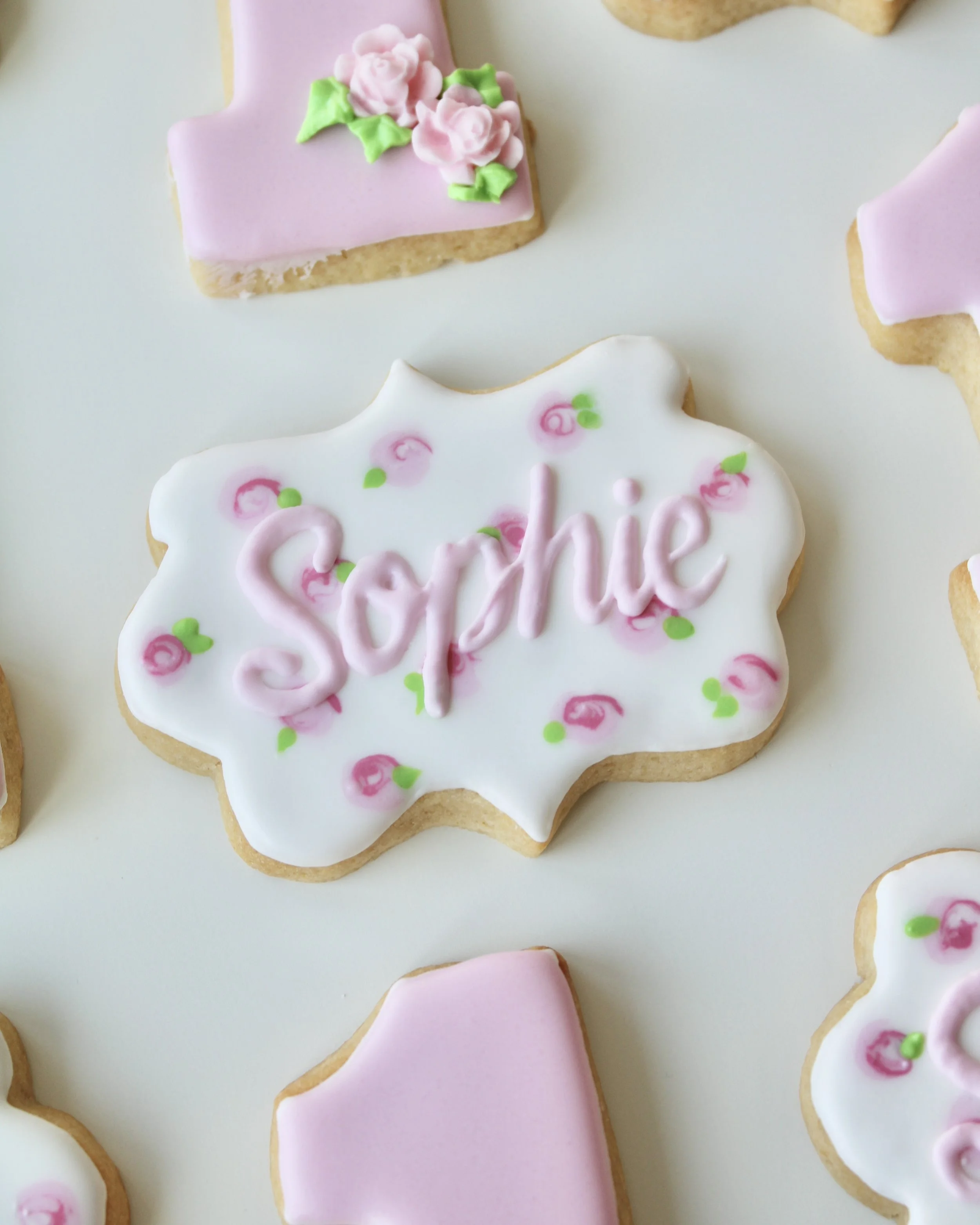 Close-up white plaque cookie decorated with “Sophie” in pink script and surrounded by hand-piped rose accents.