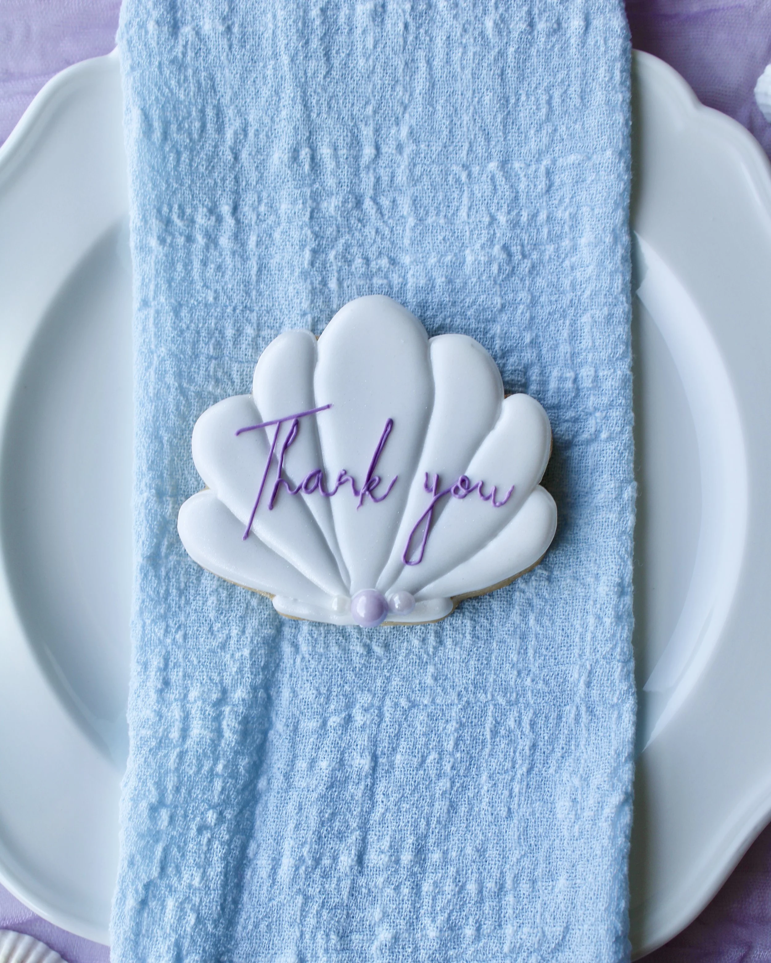 White seashell cookie with “thank you” written in purple cursive icing, placed on a white vintage plate over blue linen napkin and purple crinkled fabric with scattered seashells.