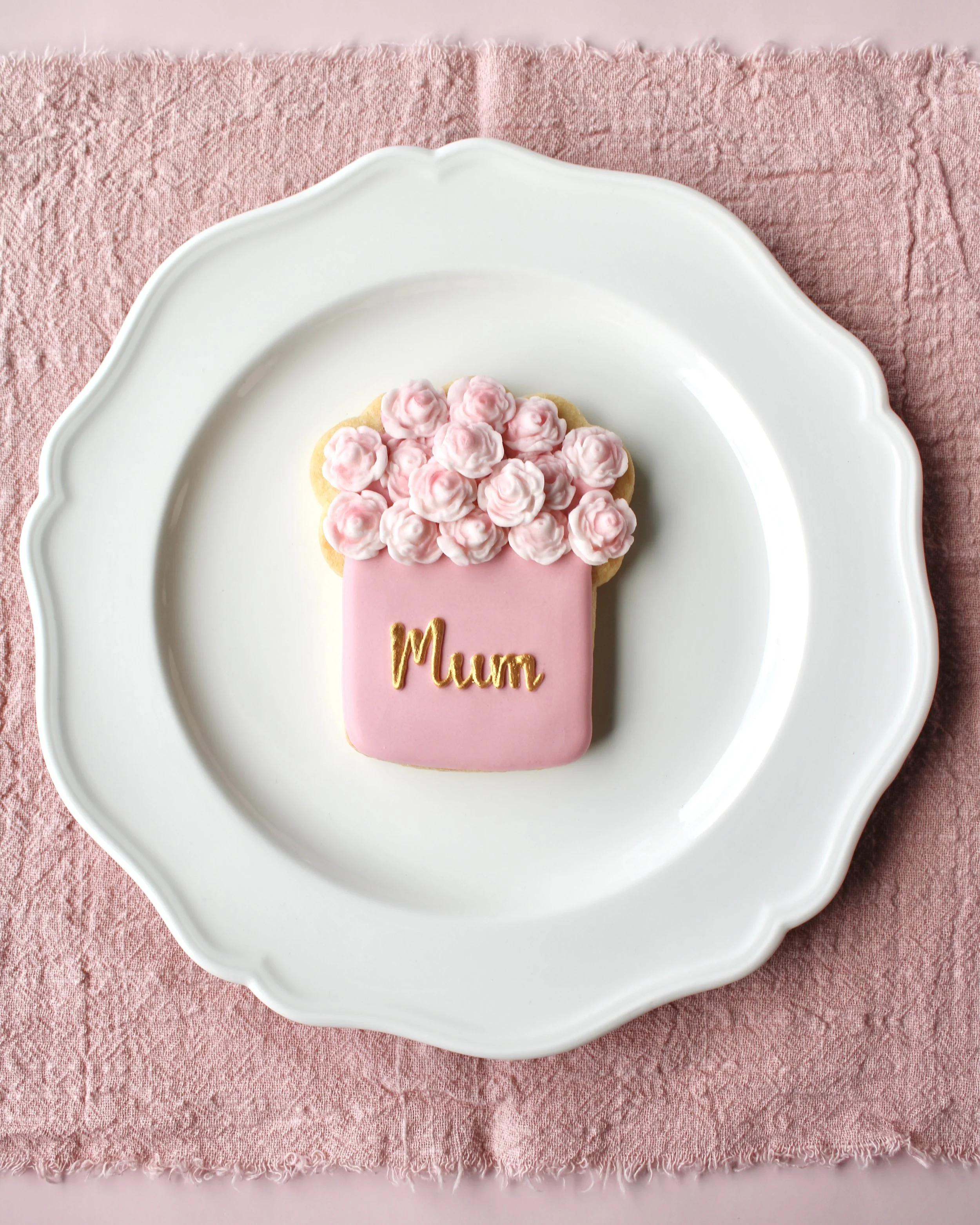 A white ornate plate with a pink flower box cookie decorated with pink roses and a gold letter 'Mum' on it, placed on a pink textured cloth.