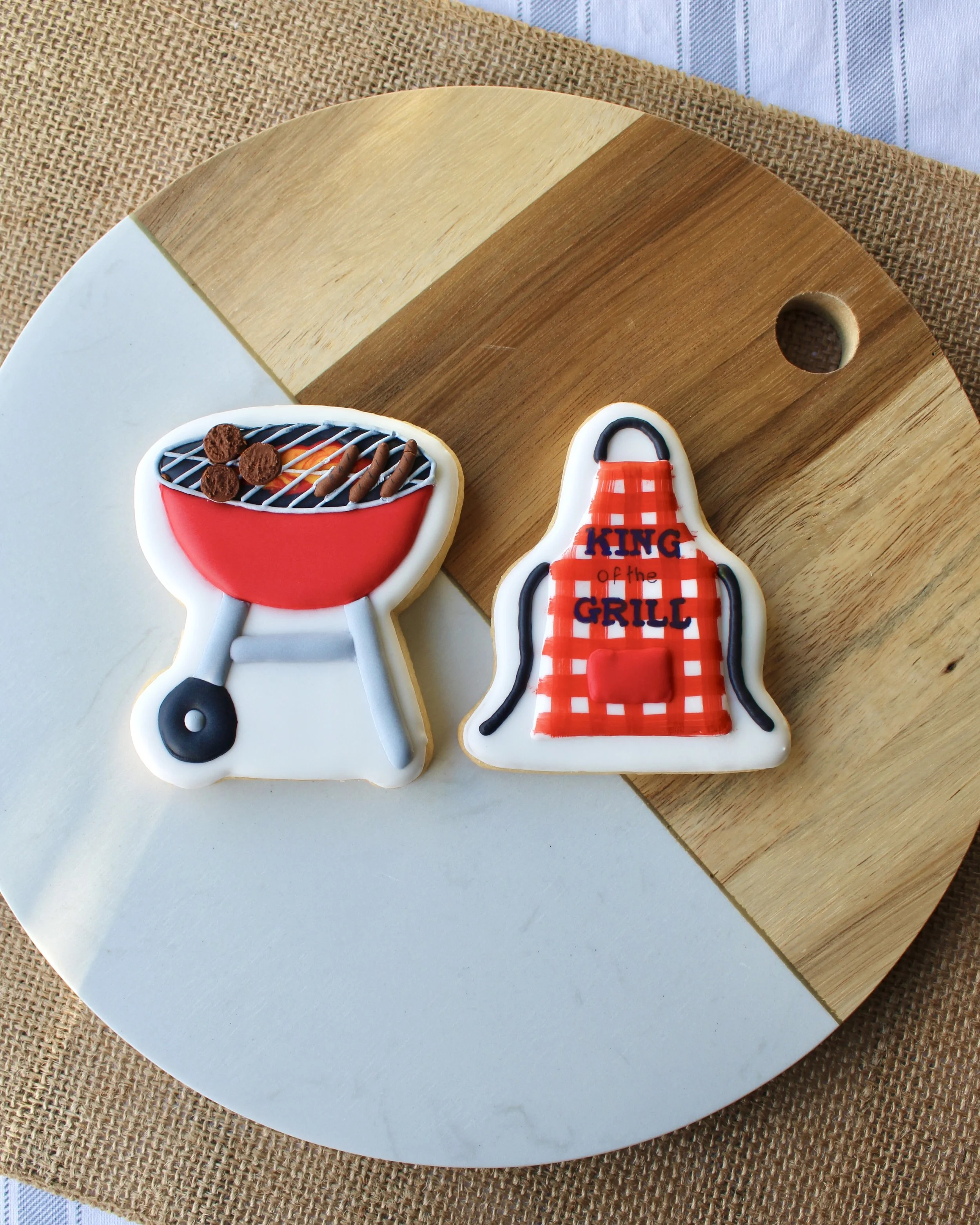 Father's Day King of the Grill cookie gift pack featuring red BBQ and gingham apron cookies with "King of the Grill" text, styled on a two-tone wooden board and hessian fabric.