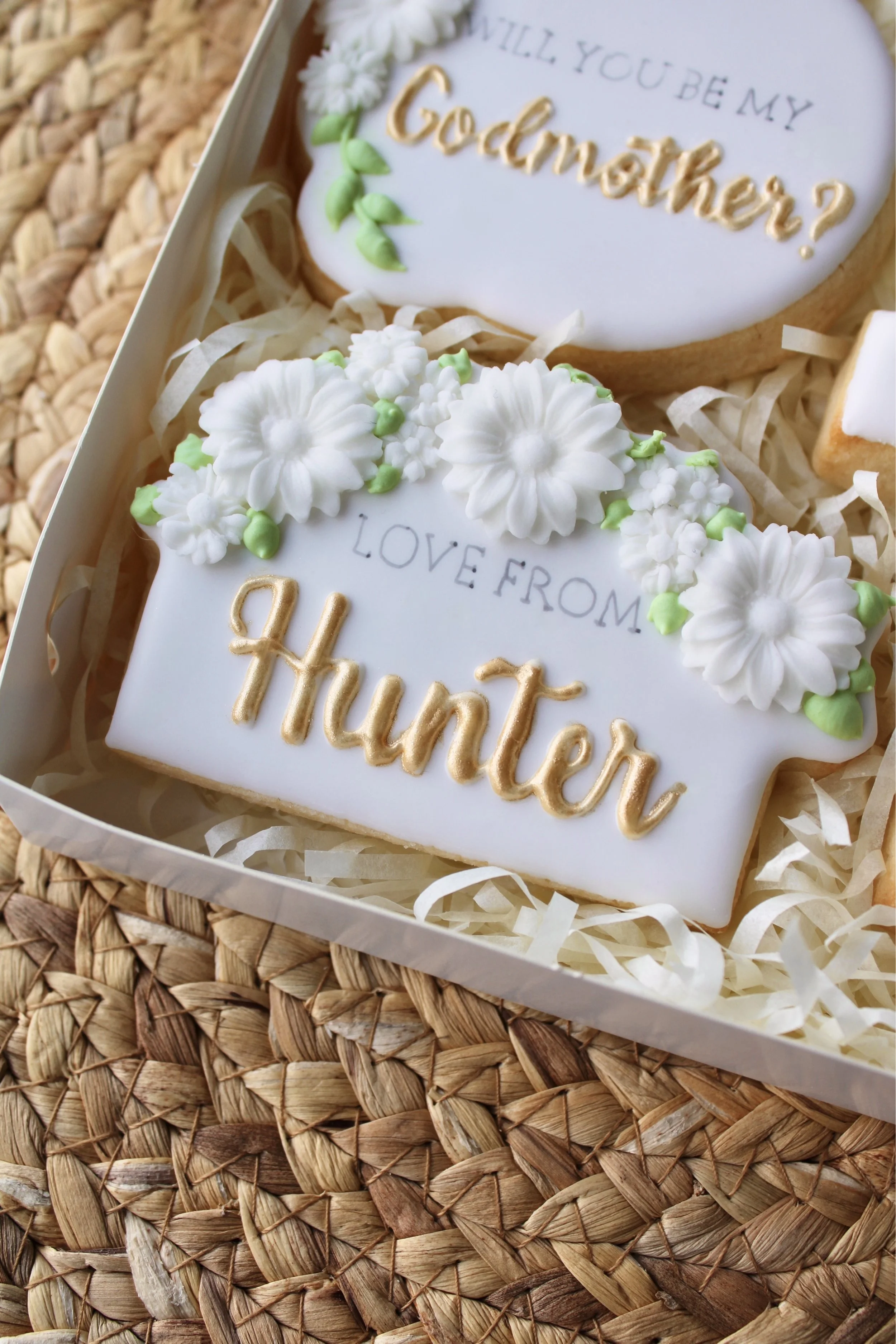 Close-up of a rectangular white iced cookie reading “Love From Hunter” with delicate leaf and white flower decorations.