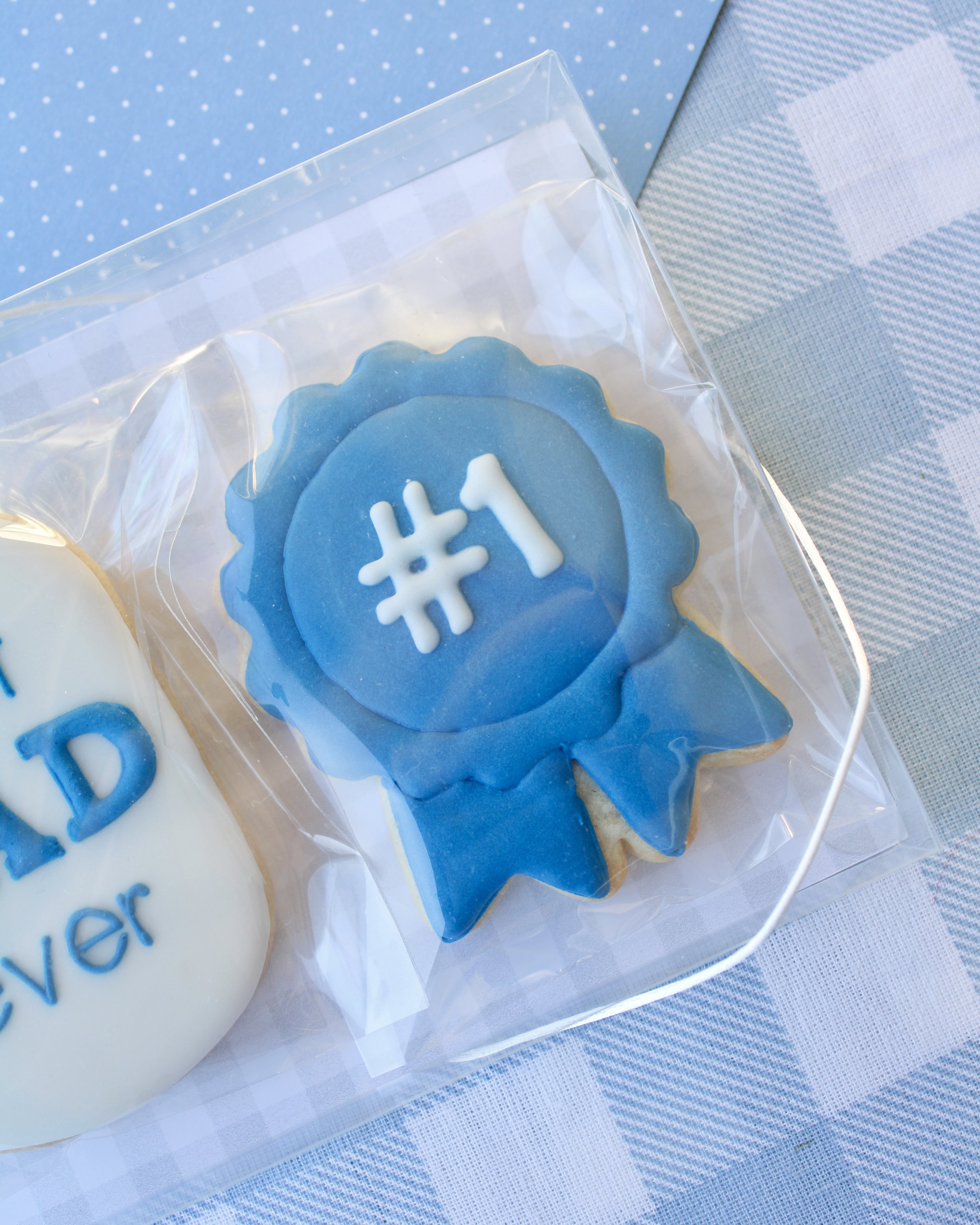 Close up of Father's Day cookie featuring a blue award ribbon cookie with "#1" in white icing, styled on blue and white gingham fabric.