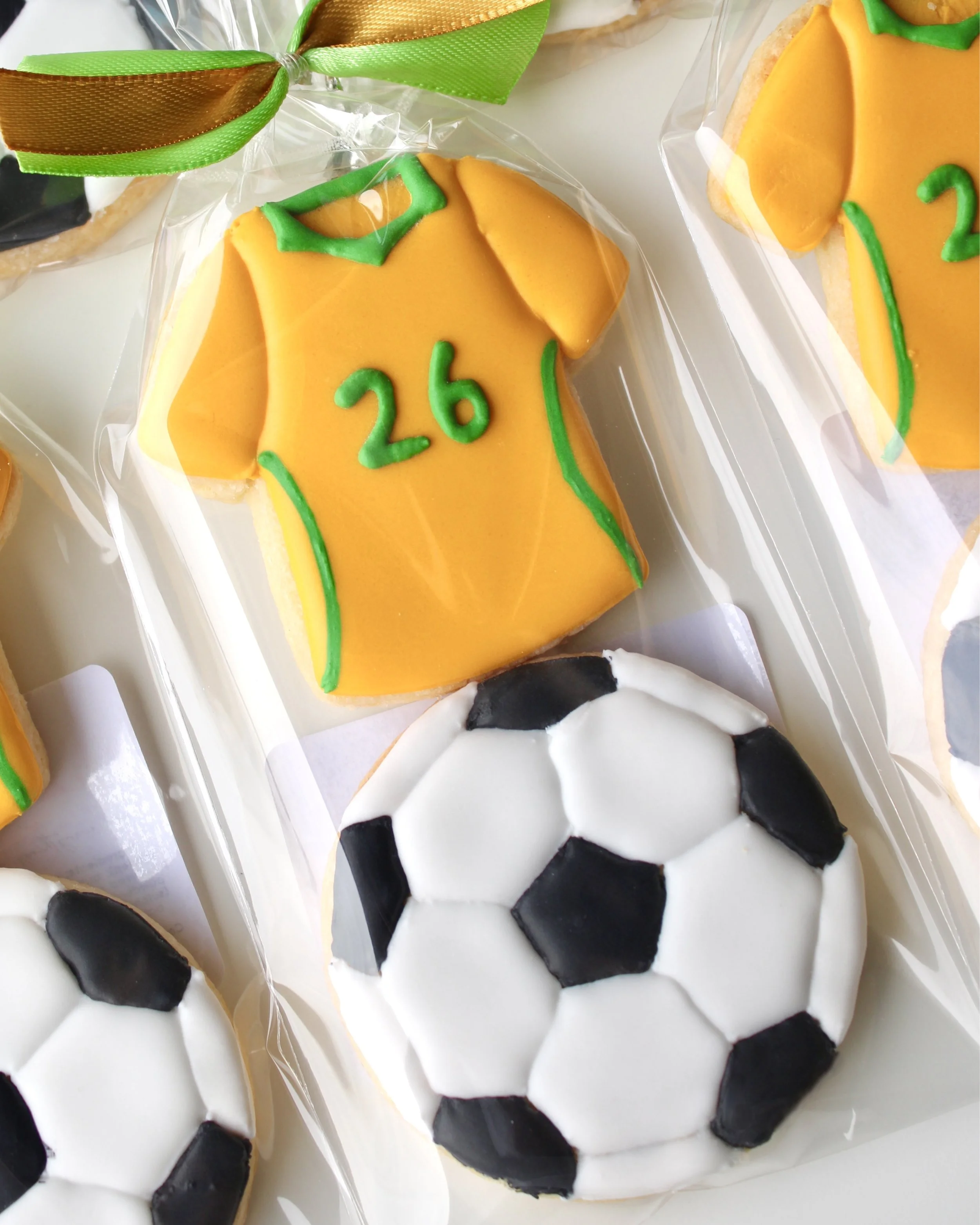 Close up of cookie pack with two mini cookies: Socceroos style jersey cookie with green accents and 26 detail, and soccer ball cookie in clear bag with ribbon.