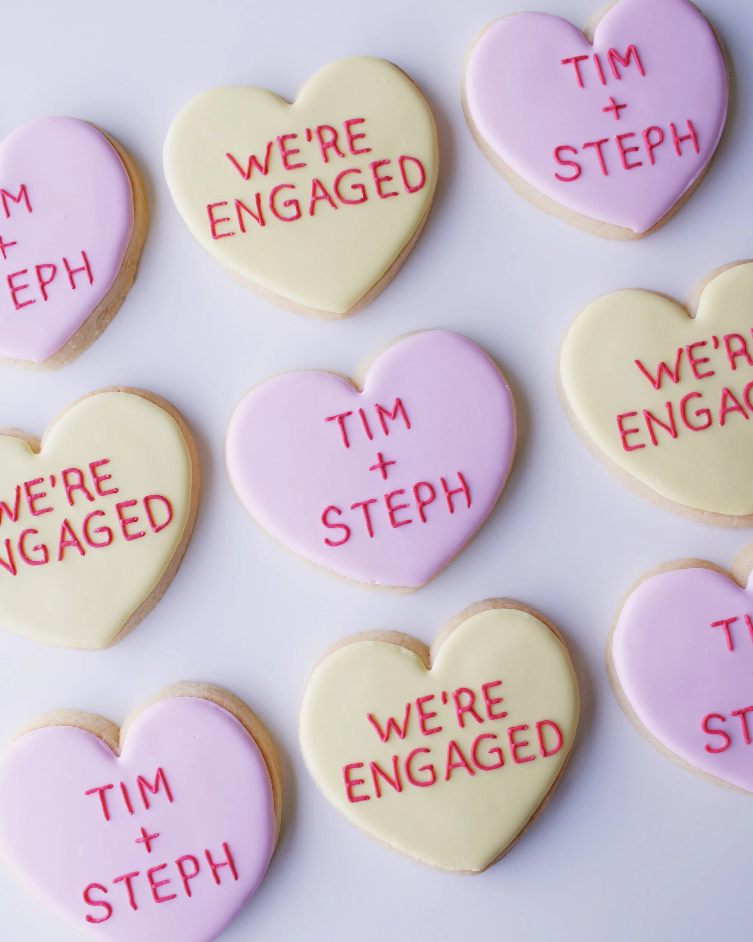 Heart-shaped cookies decorated like conversation heart candies in pastel pink and yellow icing, with red text messages such as “Tim + Steph” and “we’re engaged.”