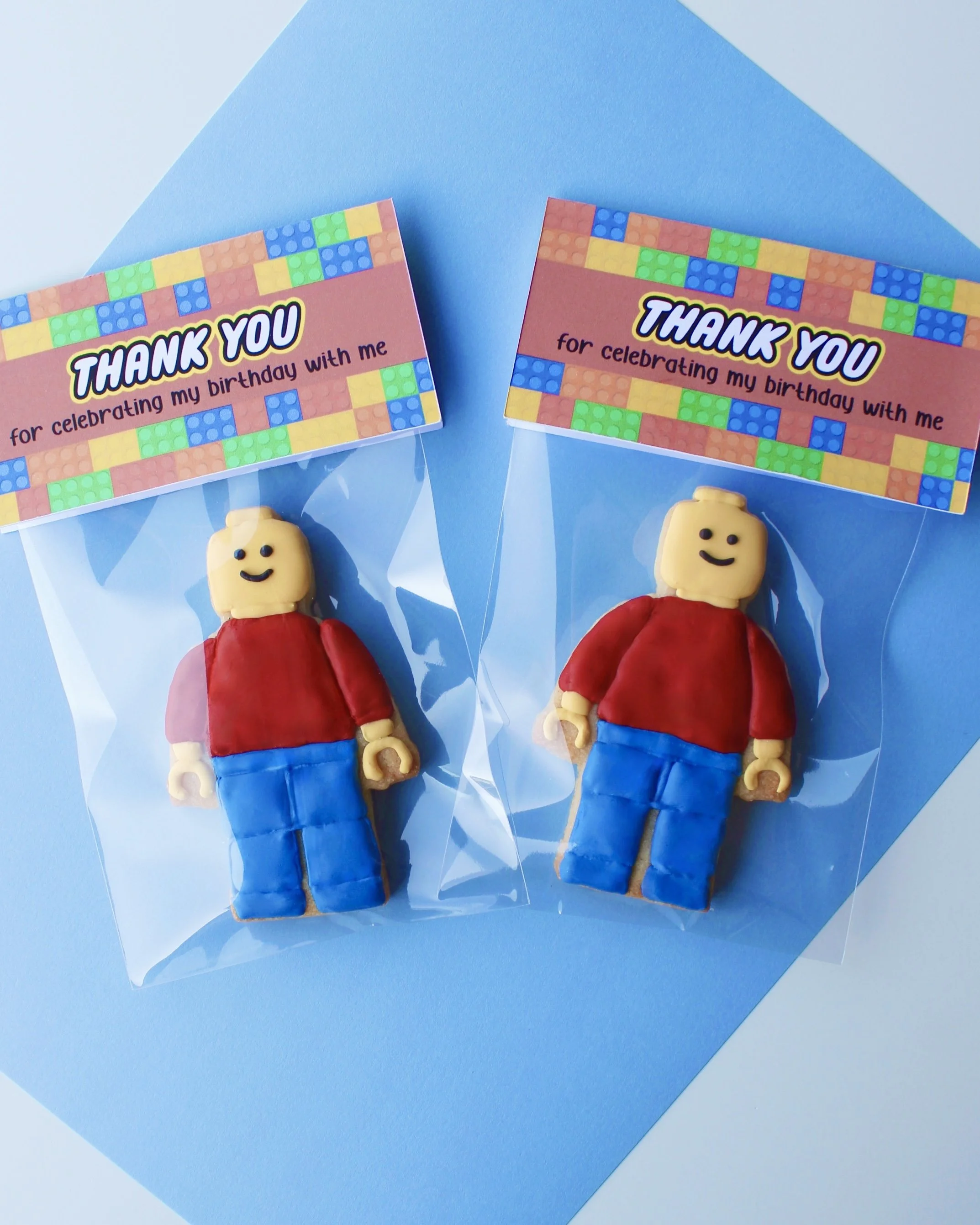 Two Lego man cookies decorated with yellow, red, and blue icing, packaged in clear cookie bags with “Thank You for Celebrating With Me” toppers and colourful building blocks in the background.