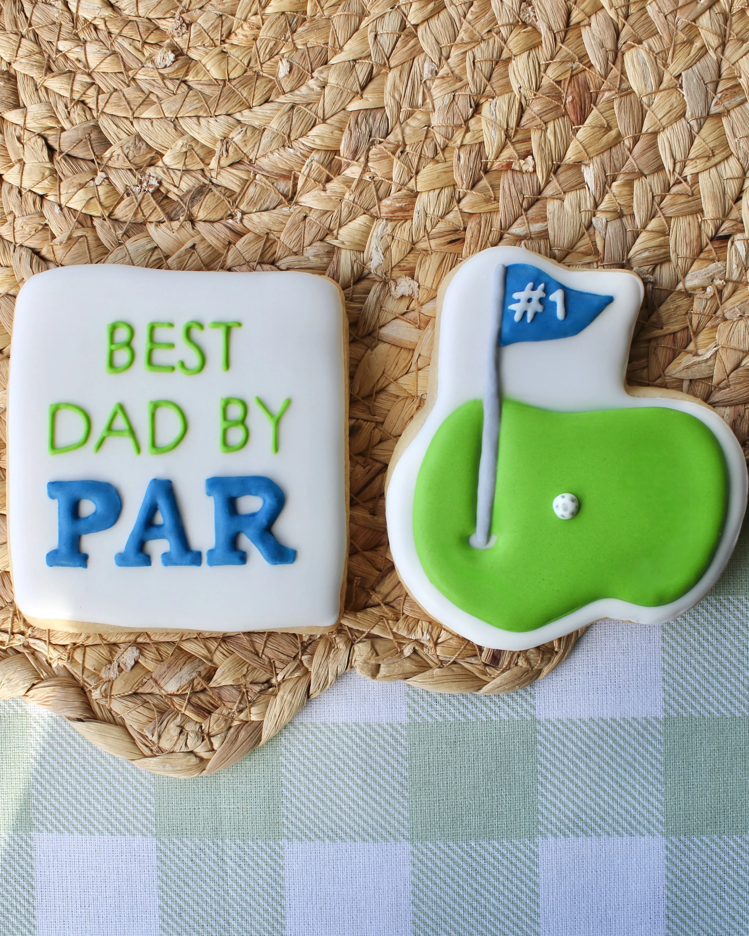 Golf-themed cookie duo set featuring a “Best Dad By Par” cookie in white with green and blue icing lettering, plus a golf par cookie with green course design and blue flag marked “#1”.