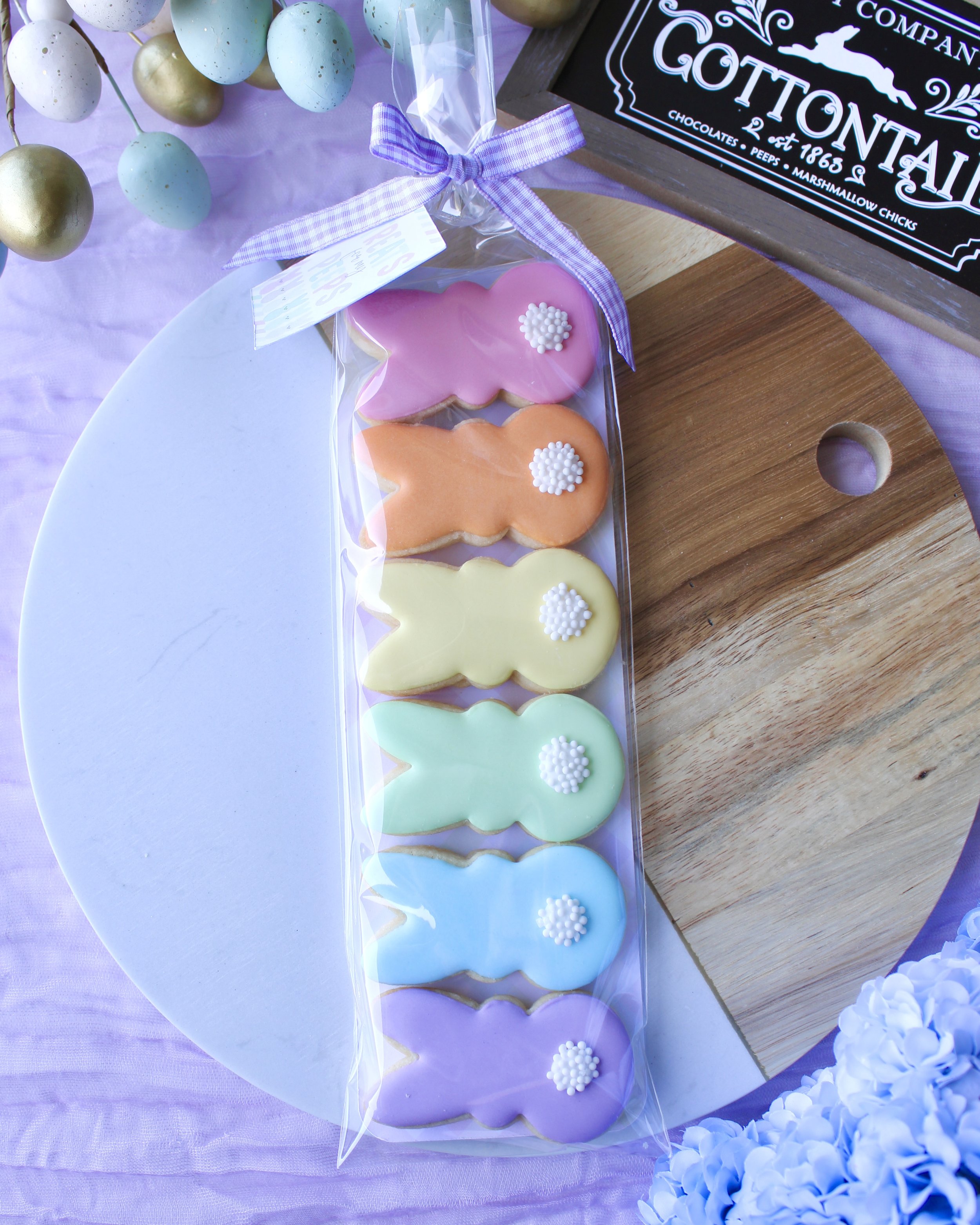 Six mini Easter Bunny Peeps cookies in pastel colours with white cotton tails in a clear bag tied with gingham bow, styled on marble and wood board with lilac fabric, eggs and hydrangeas.