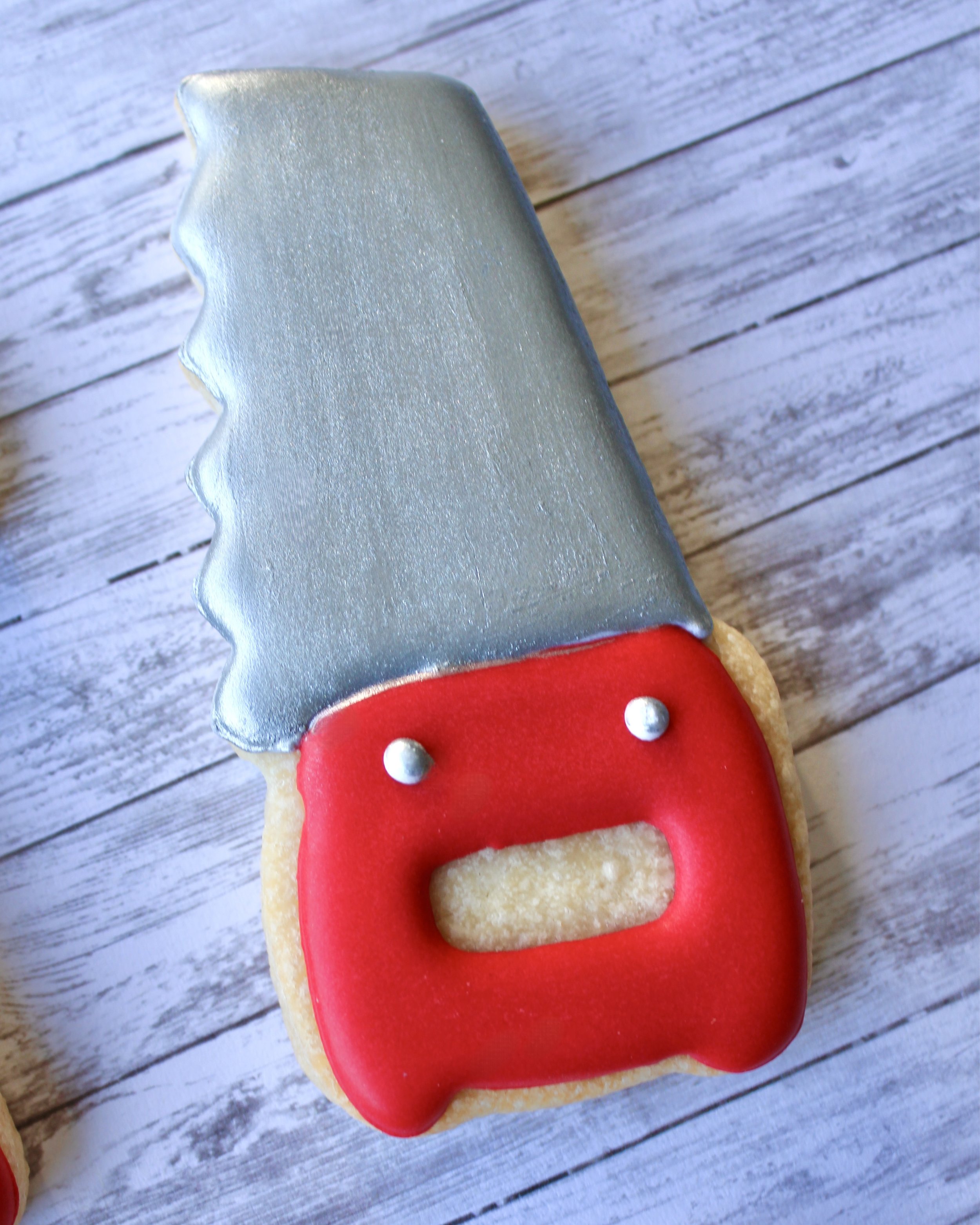Close up of Father's Day tool cookie shaped like a saw, with silver icing and a red icing handle, styled on a grey washed wooden background.