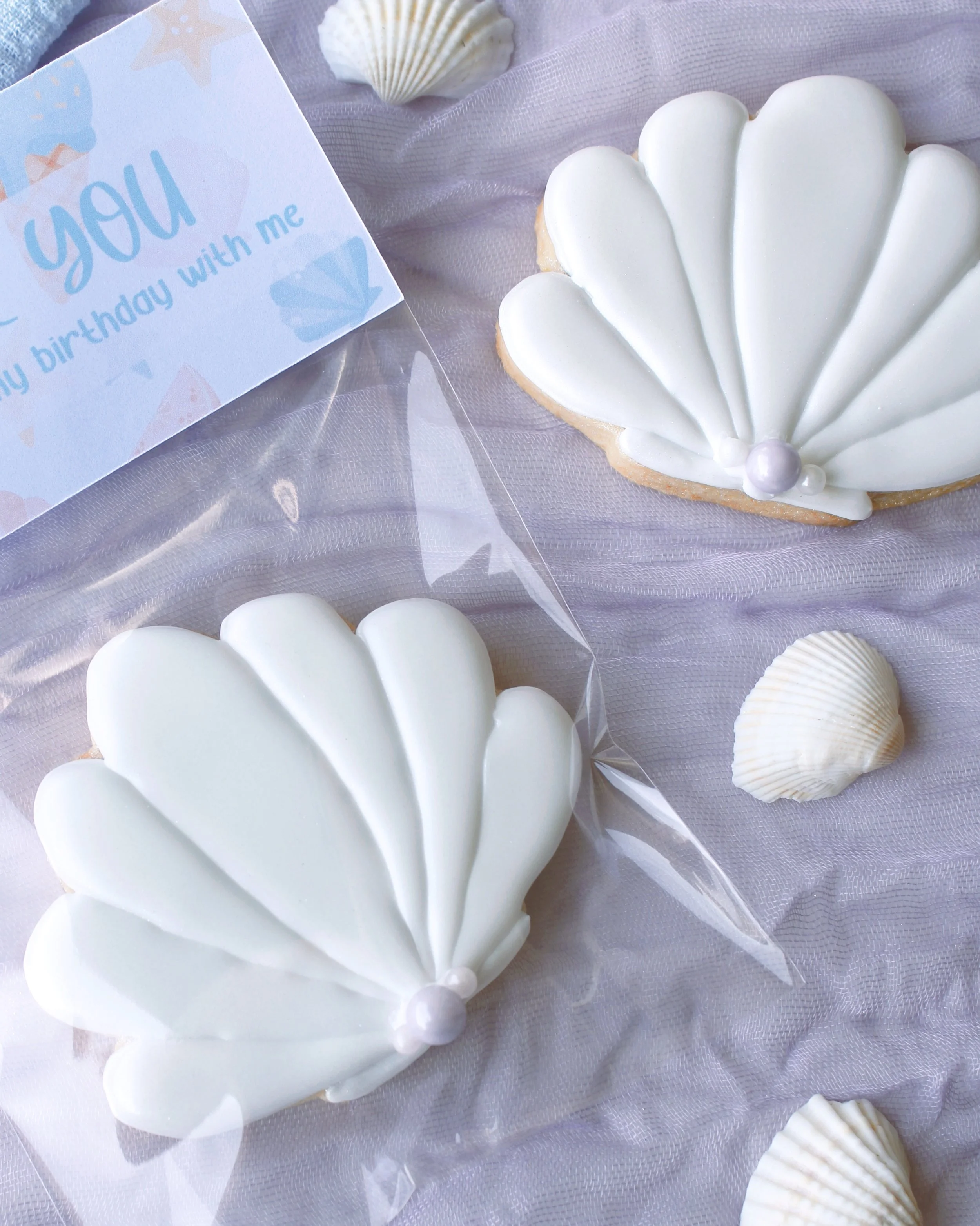 Seashell birthday party favour cookie in shimmering white with pearl details, packaged with a mermaid-themed “thank you for celebrating my birthday with me” topper on purple crinkled fabric.