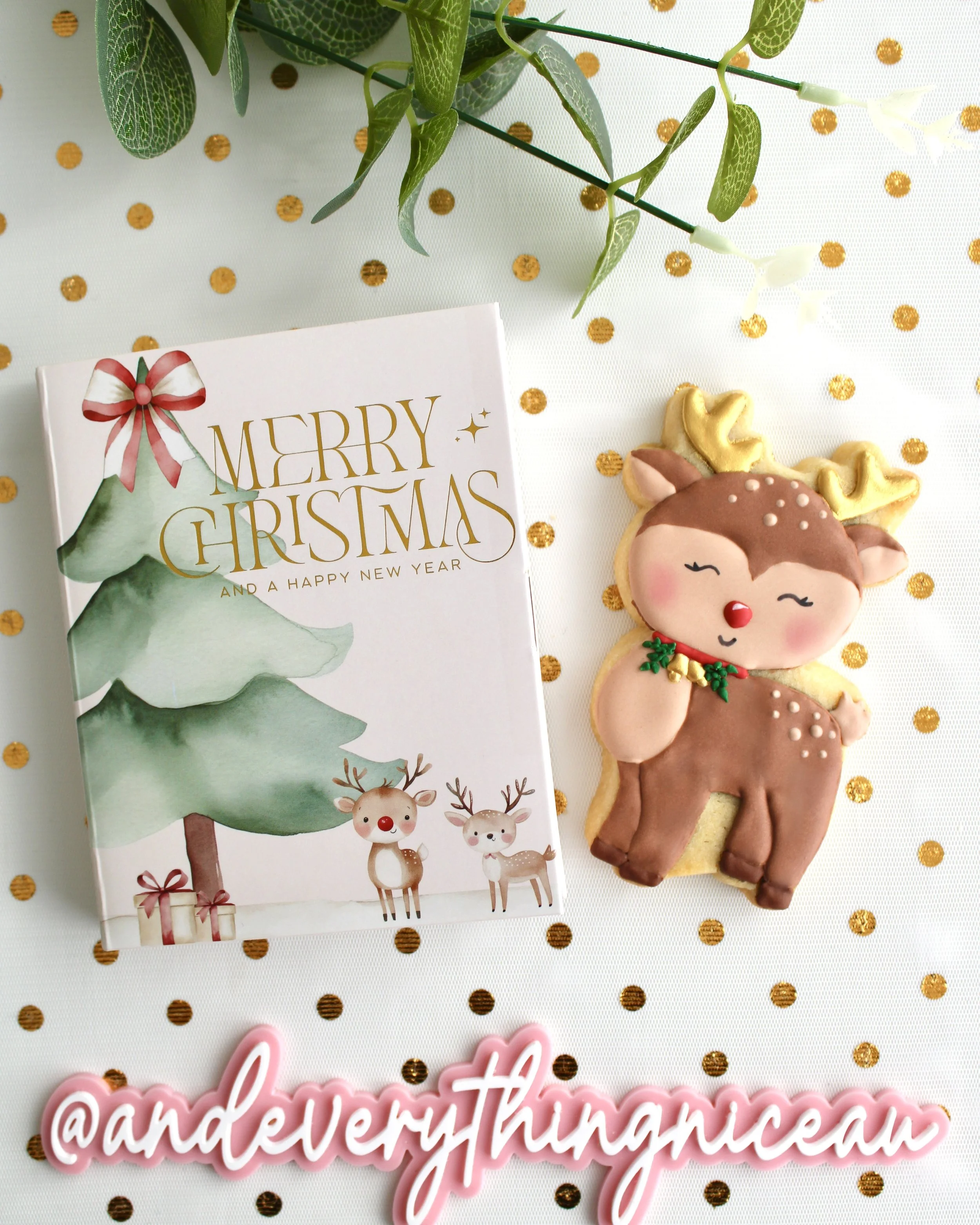 A festive Christmas cookie card box with watercolour trees and reindeer sits beside a brown and tan reindeer cookie with gold accents, styled on white sheer fabric with gold dots and a pink “@andeverythingnice” watermark plaque.