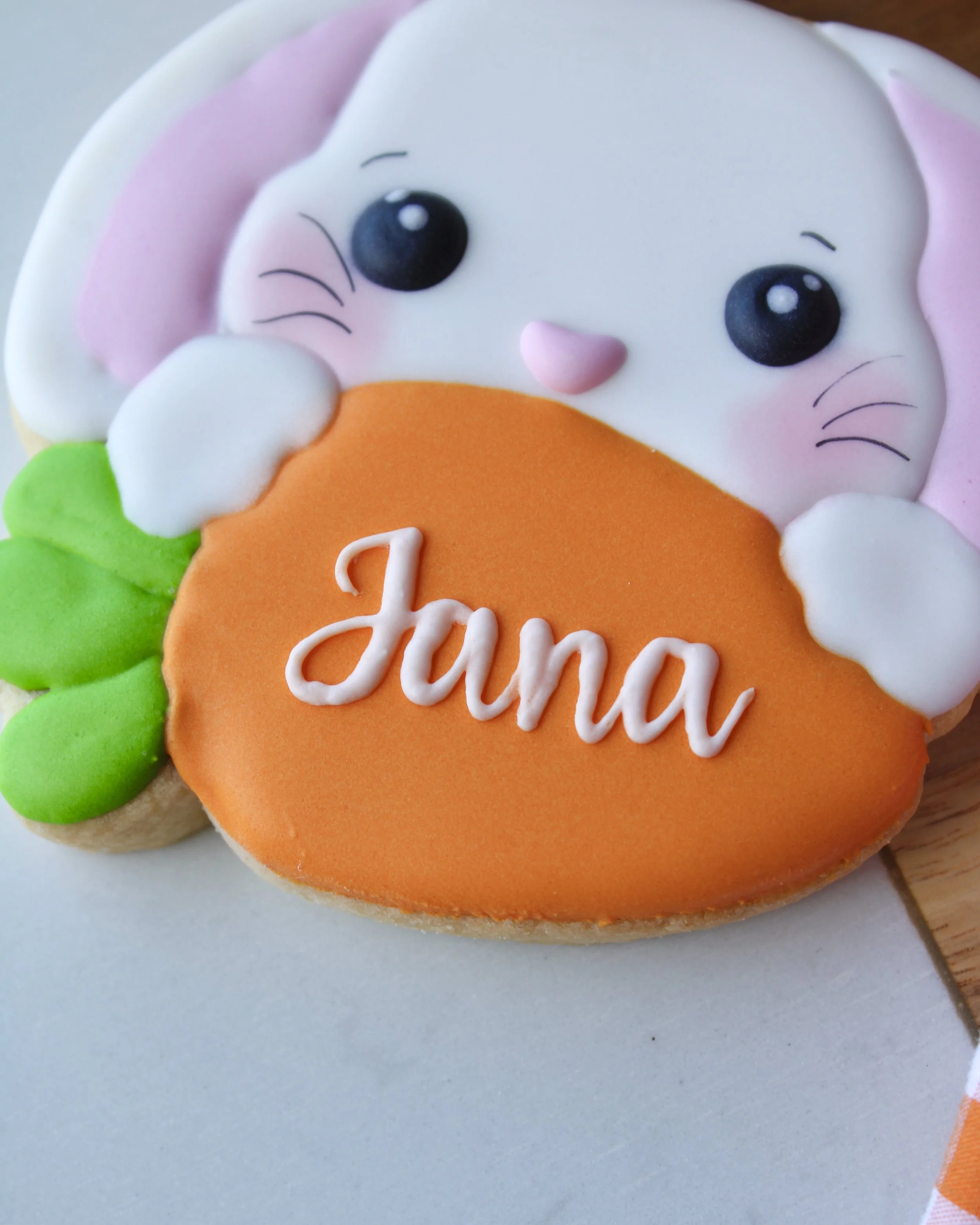 Close-up personalised Easter bunny sugar cookie with white face, pink ears and nose, holding an orange and green carrot with “Jana” in white script icing, styled on half wood and half marble board.