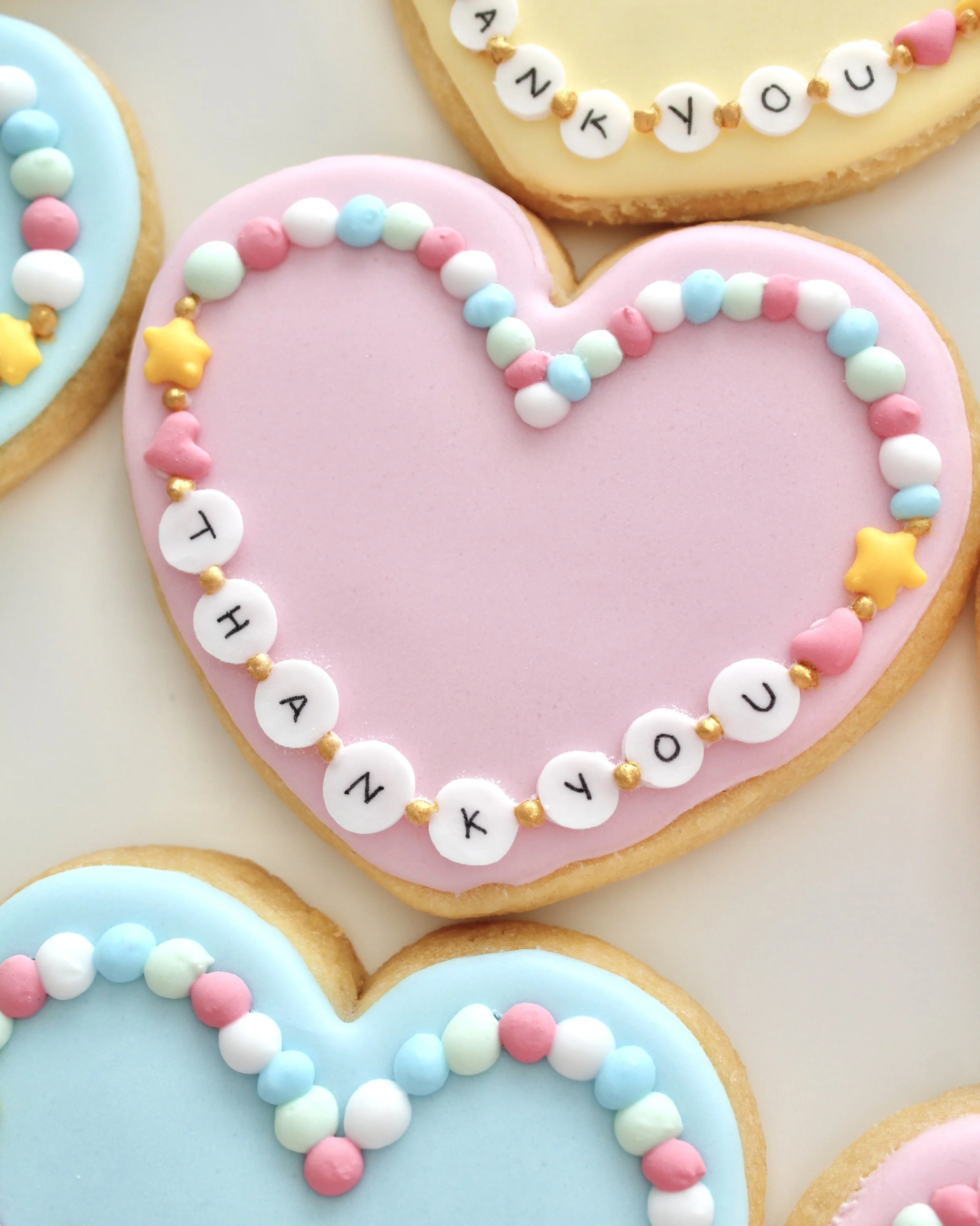 Close-up of a pastel pink heart-shaped cookie decorated like a friendship bracelet, featuring “thank you” on the icing beads and additional beads in soft pastel colours.