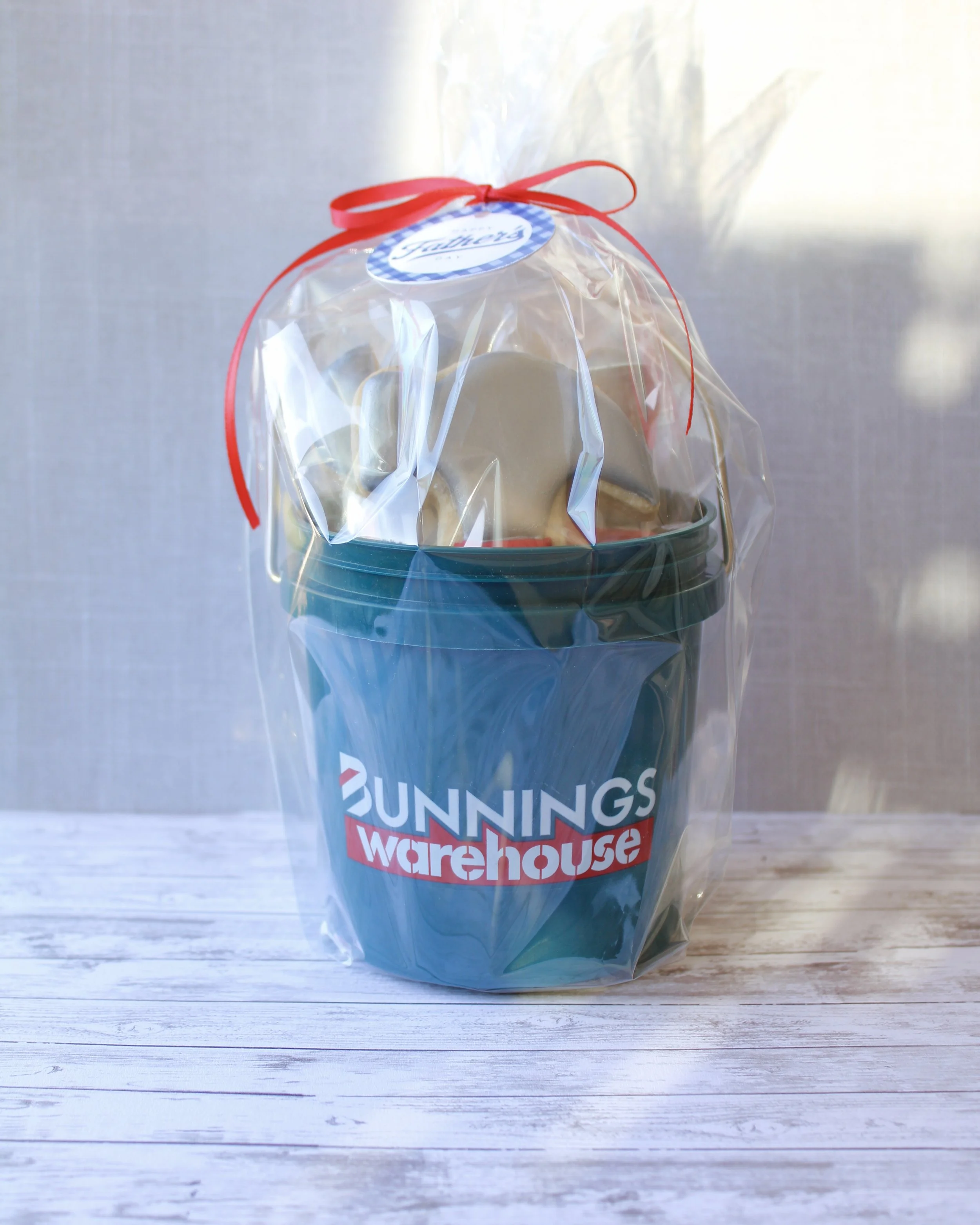 Father's Day tool cookies in a dark green Bunnings Warehouse mini bucket, wrapped in cellophane with a red ribbon and Happy Father's Day gift tag on a grey washed wooden background.