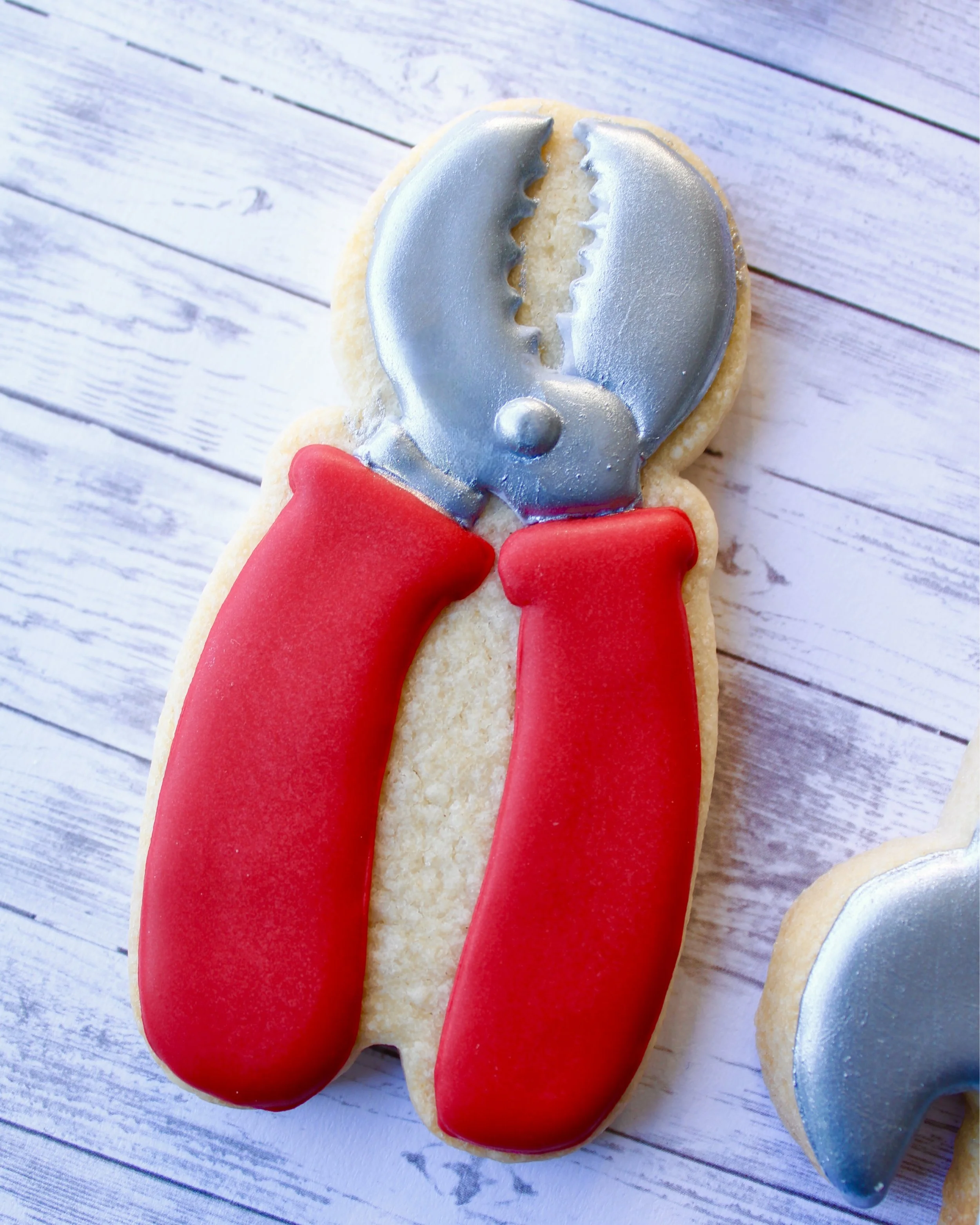 Close up of Father's Day tool cookie shaped like a pair of pliers, with silver icing and a red icing handle, styled on a grey washed wooden background.