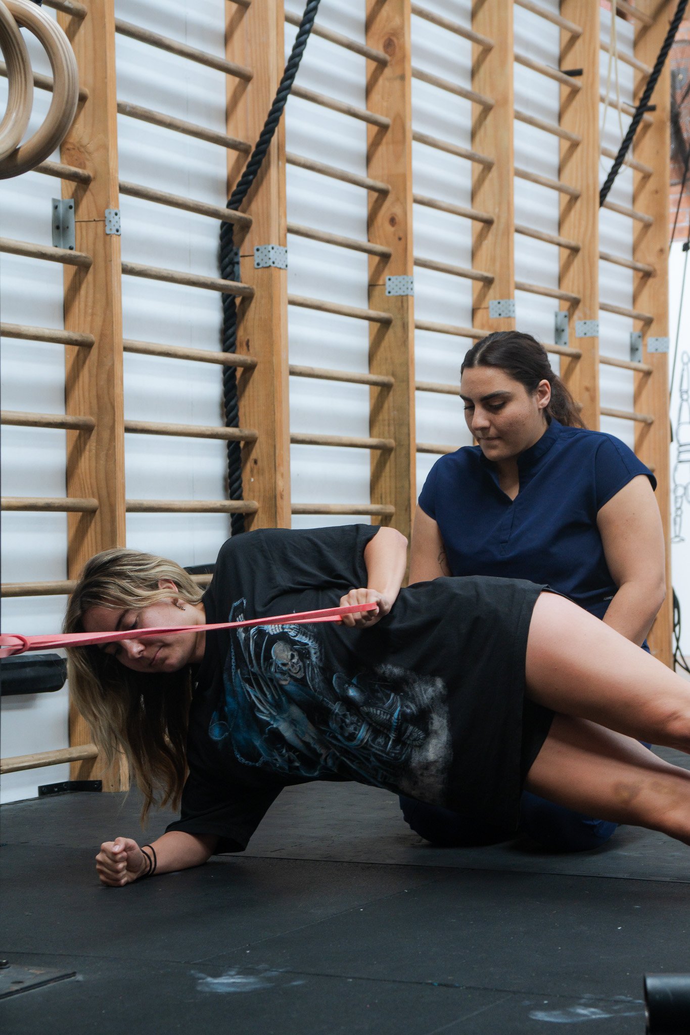 A woman is doing a resistance band exercise on the floor, assisted by another woman in a gym or training room with wooden wall bars and gym equipment.