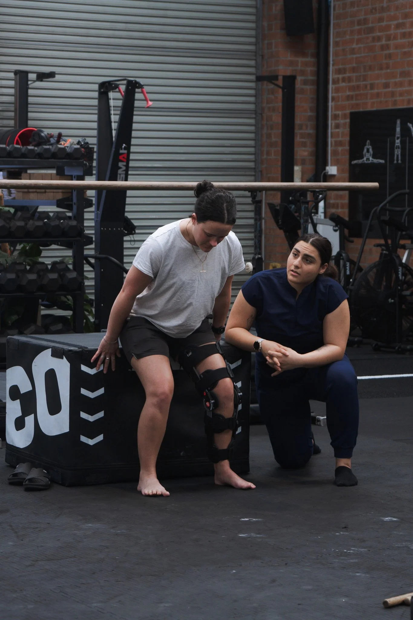 A woman with a brace on her knee, sitting on a workout box, appears to be injured or in pain, assisted by a woman kneeling beside her in a gym setting.