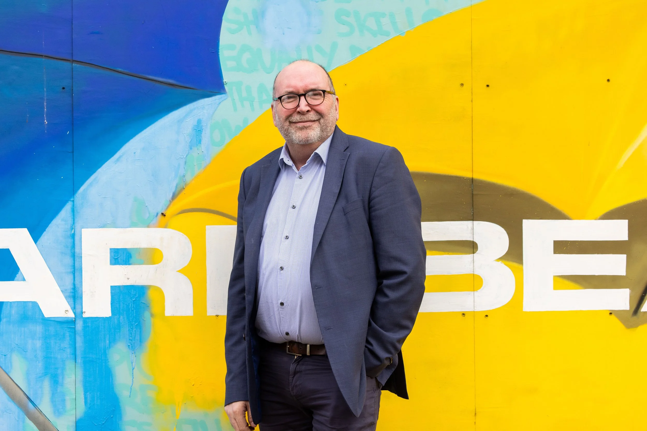 A smiling man in a gray suit, light blue shirt, and glasses standing in front of a colorful mural with blue, yellow, and green sections and white text.