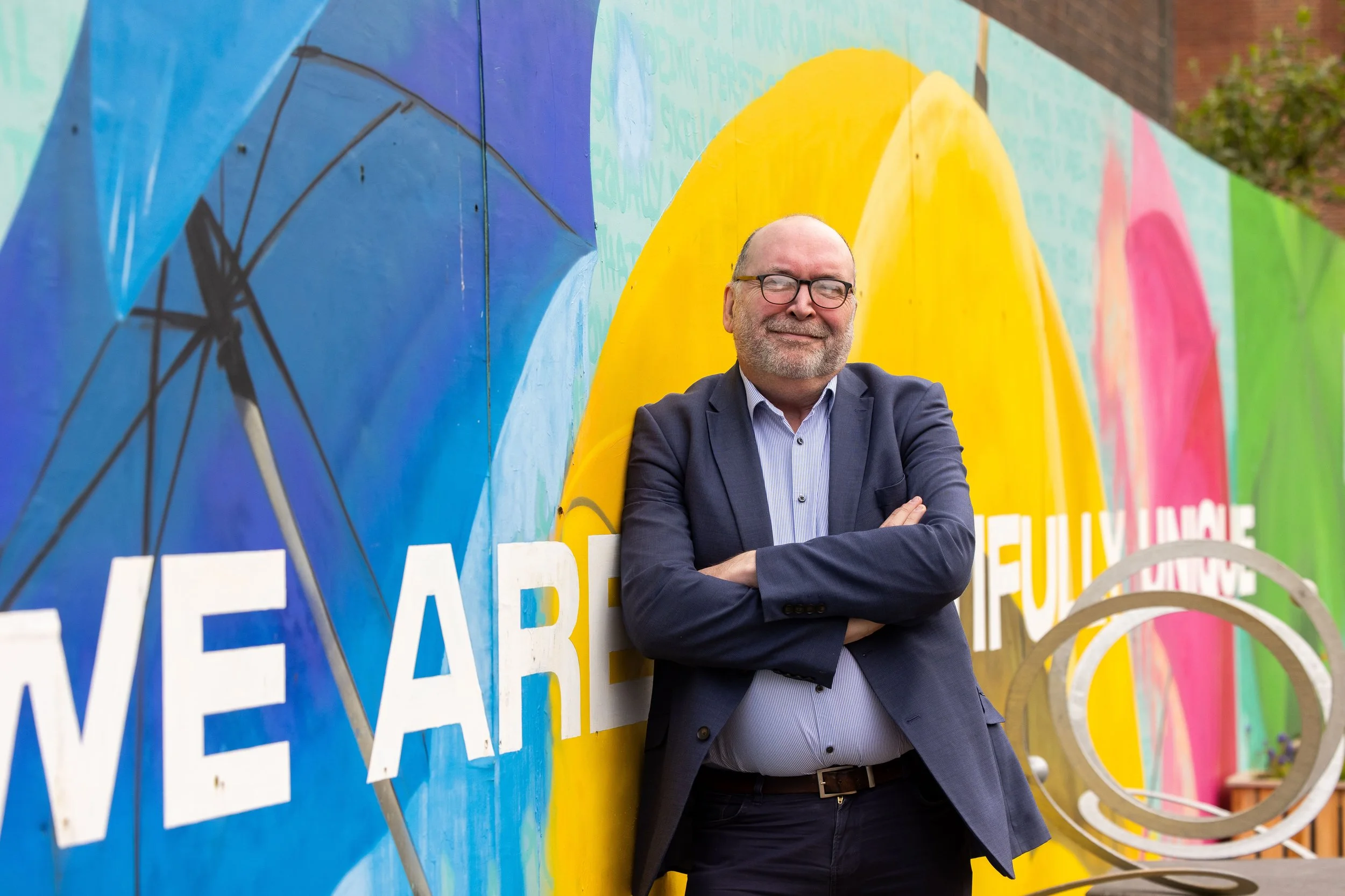 A man in a blue suit with glasses standing with crossed arms, smiling, in front of a colorful mural with large text and abstract designs.