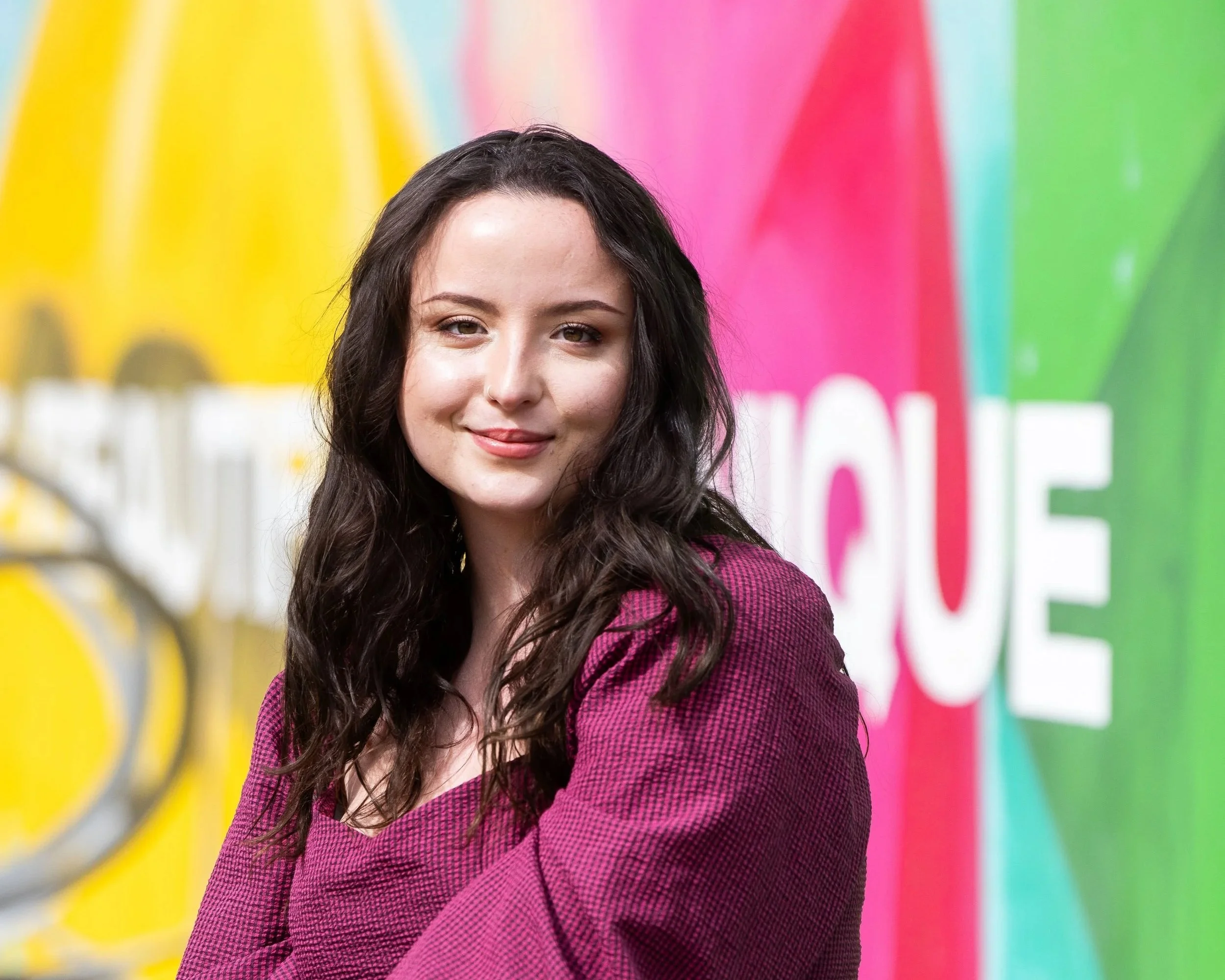 A young woman with long dark wavy hair and light skin, smiling, standing in front of a colorful background with yellow, pink, and green sections.