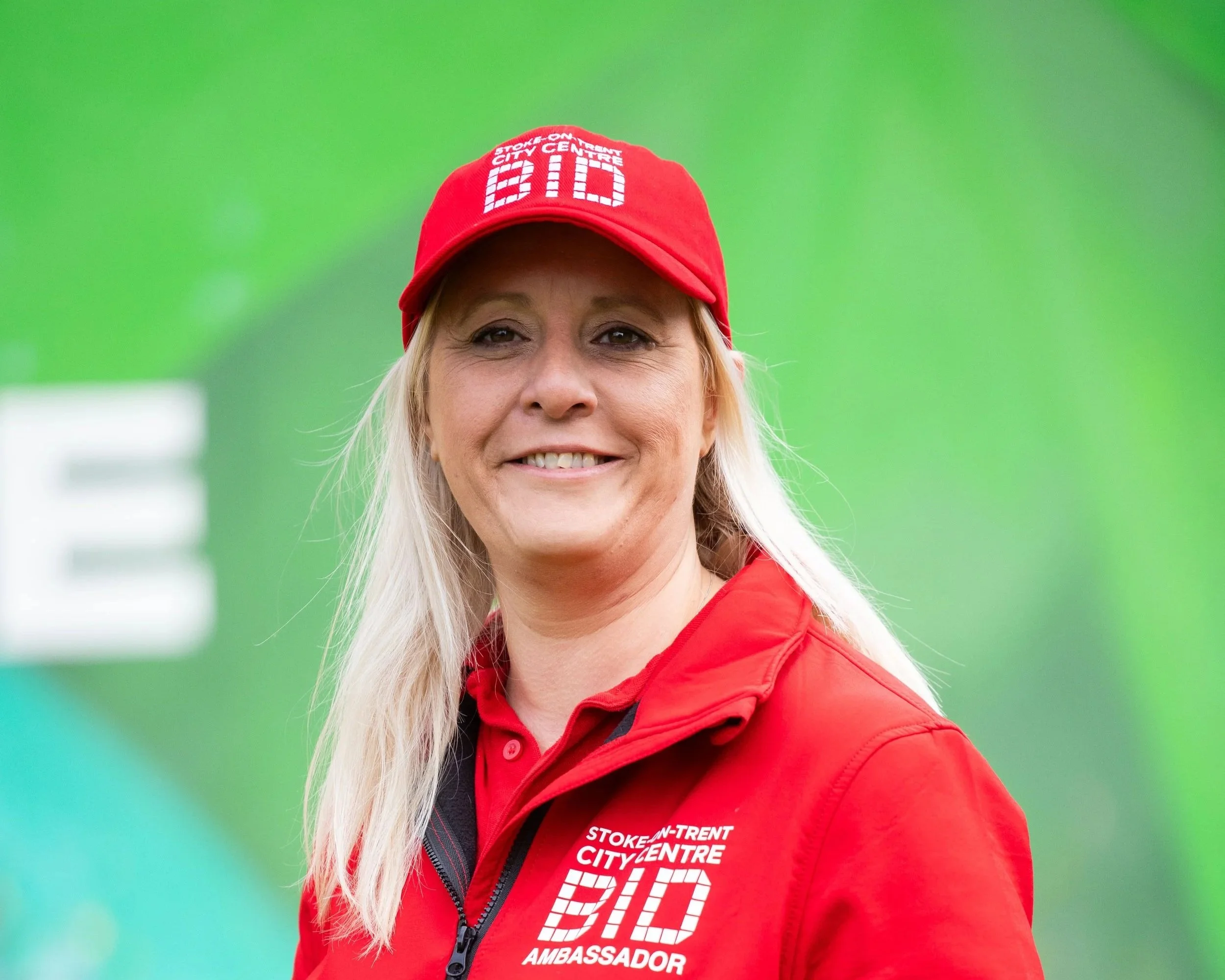 A woman smiling wearing a red jacket and red cap with white text, standing outdoors with a green background.