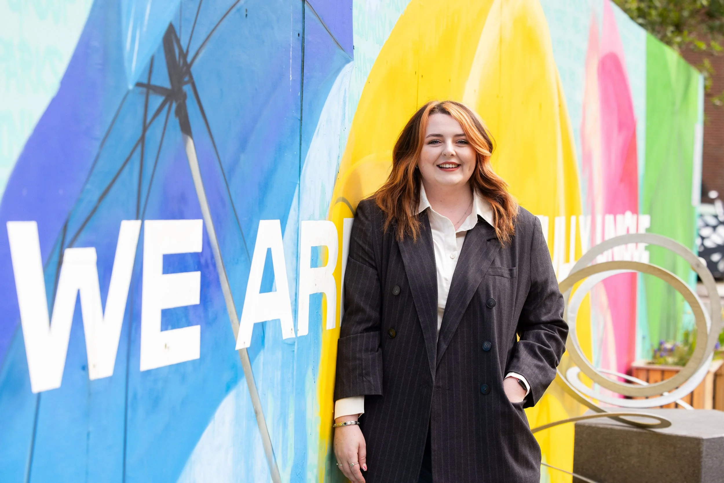 A young woman with red hair smiling and standing in front of a colorful mural with large text and rainbow rainbow patterns. She is wearing a dark blazer over a white shirt.