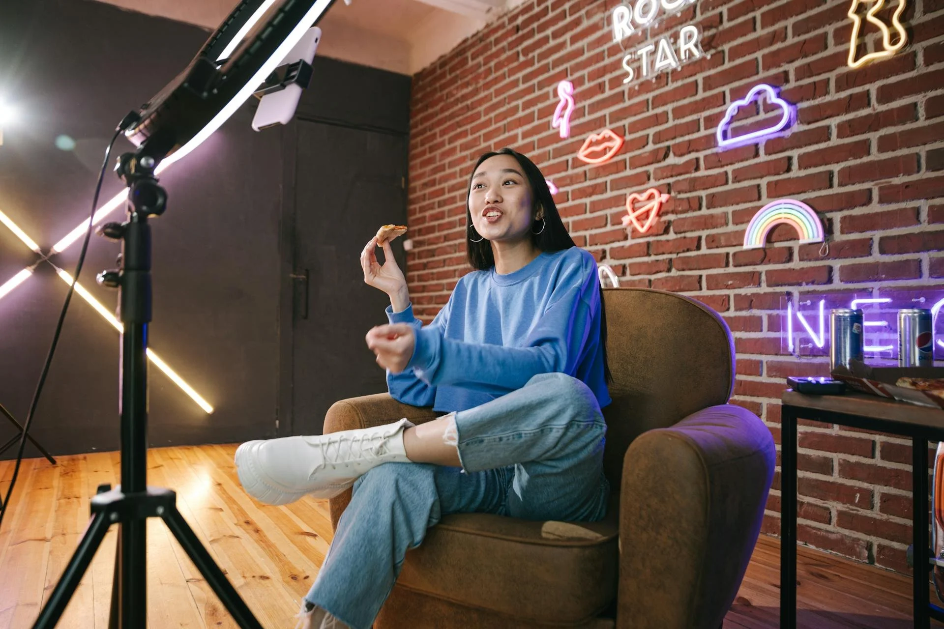 A young woman sitting on a brown armchair in a room with brick walls, neon signs, and a wooden floor, recording a video with a smartphone mounted on a tripod, with snacks and soda cans on a small table beside her.