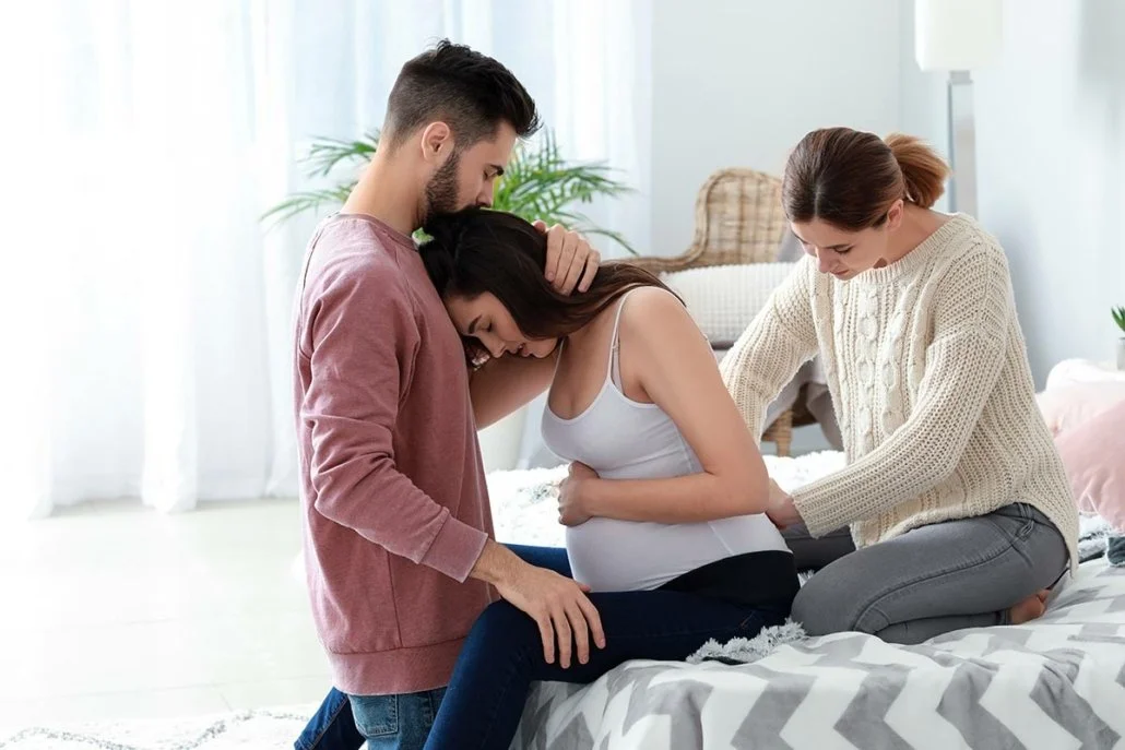 A pregnant woman is surrounded by two people, one man and one woman, in a supportive scene in a bright, airy bedroom.