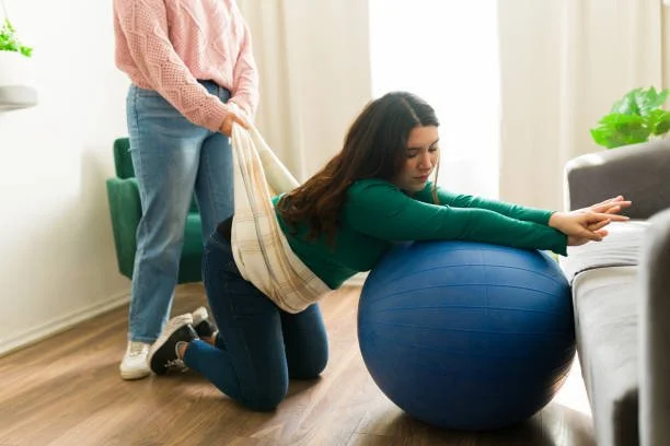A woman practicing physical therapy with a large exercise ball while a therapist assists her using a strap in a living room.