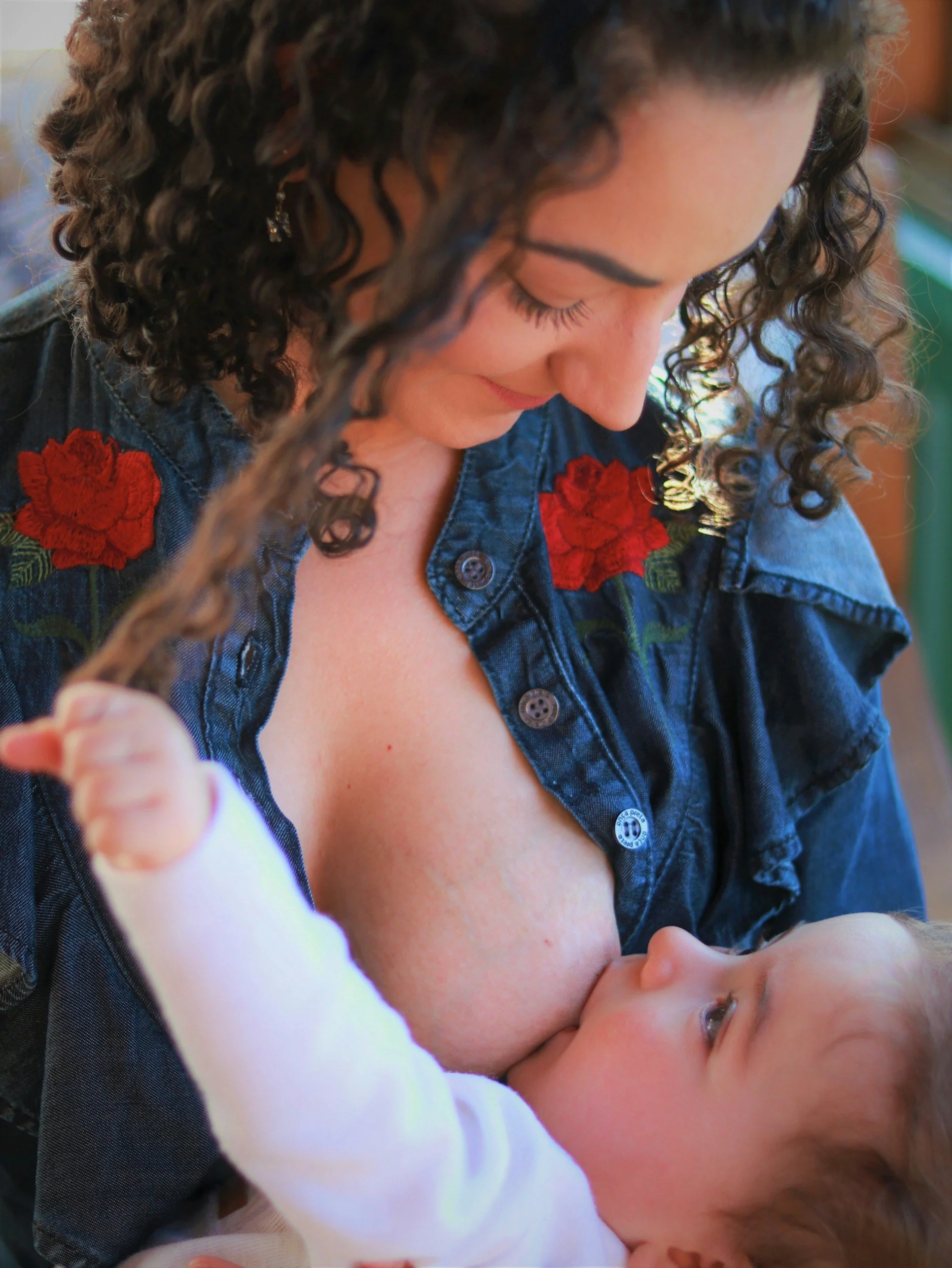 A woman with curly dark hair and floral embroidery on her denim jacket is gently kissing a young child's forehead, who is lying down and looking up with light brown hair and blue eyes.