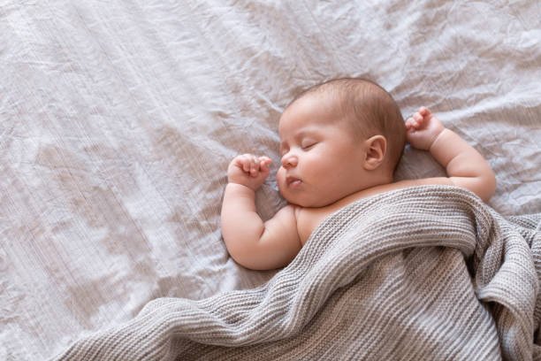A sleeping baby lying on a bed, covered with a beige blanket.