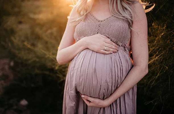 A pregnant woman standing outdoors at sunset, wearing a sleeveless top and a long skirt, holding her belly with both hands.