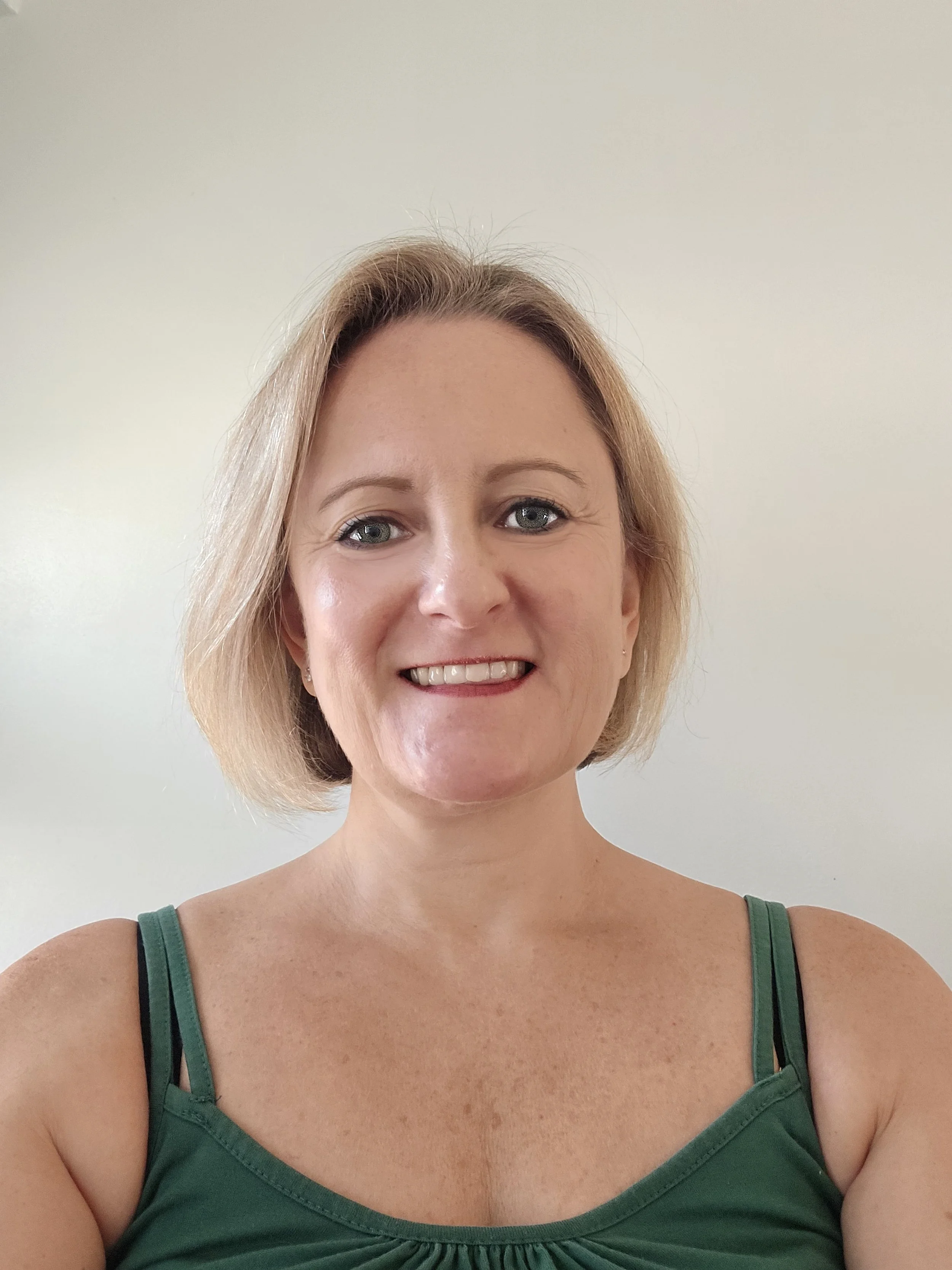 Kelly Woolhouse Birth Doula and qualified Hypnobirthing Instructor with shoulder-length blonde hair, green eyes, and a bright smile, wearing a green tank top with thin straps, posing against a plain light-coloured background.