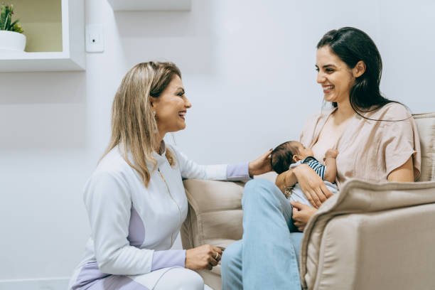 A woman holding a baby on her lap and another woman sitting beside her, smiling and engaging in conversation in a hospital room.