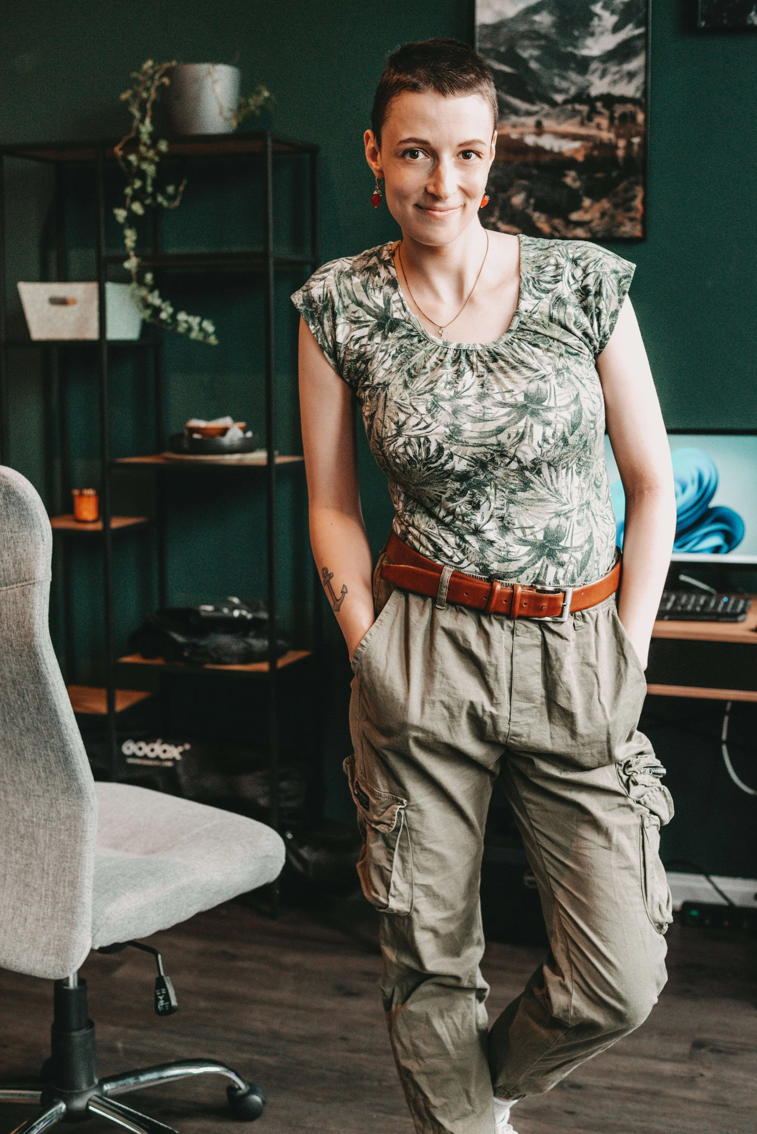 Eine Frau mit mit kurzen Haaren, buntem T-Shirt und Cargo-Hosentaschen, steht in einem modernen Raum mit dunkelgrüner Wand, einer Pflanzenvase, einem Regal.