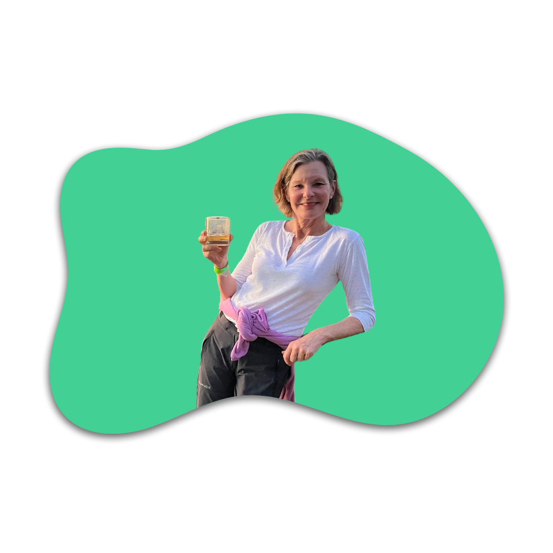 Eine lächelnde Frau hält ein Glas mit einem Getränk in der Hand, trägt einen weißen Hemd, mit einem Pullover um die Taille gebunden, vor einem grünen Hintergrund mit unregelmäßiger Form.