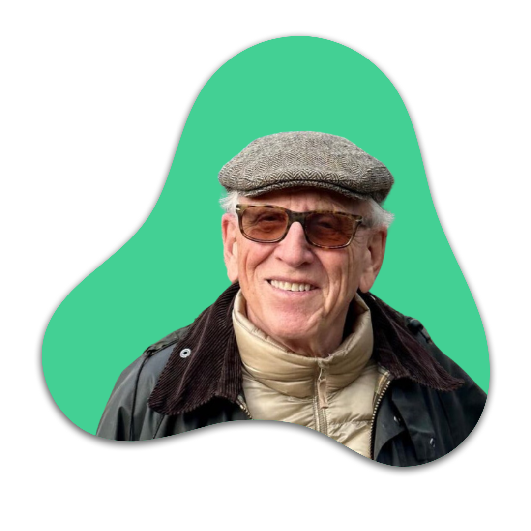 Ein älterer Mann trägt eine Schiebermütze, Sonnenbrille, eine beige Jacke und eine schwarze Jacke davor, steht vor einem grünen Hintergrund.