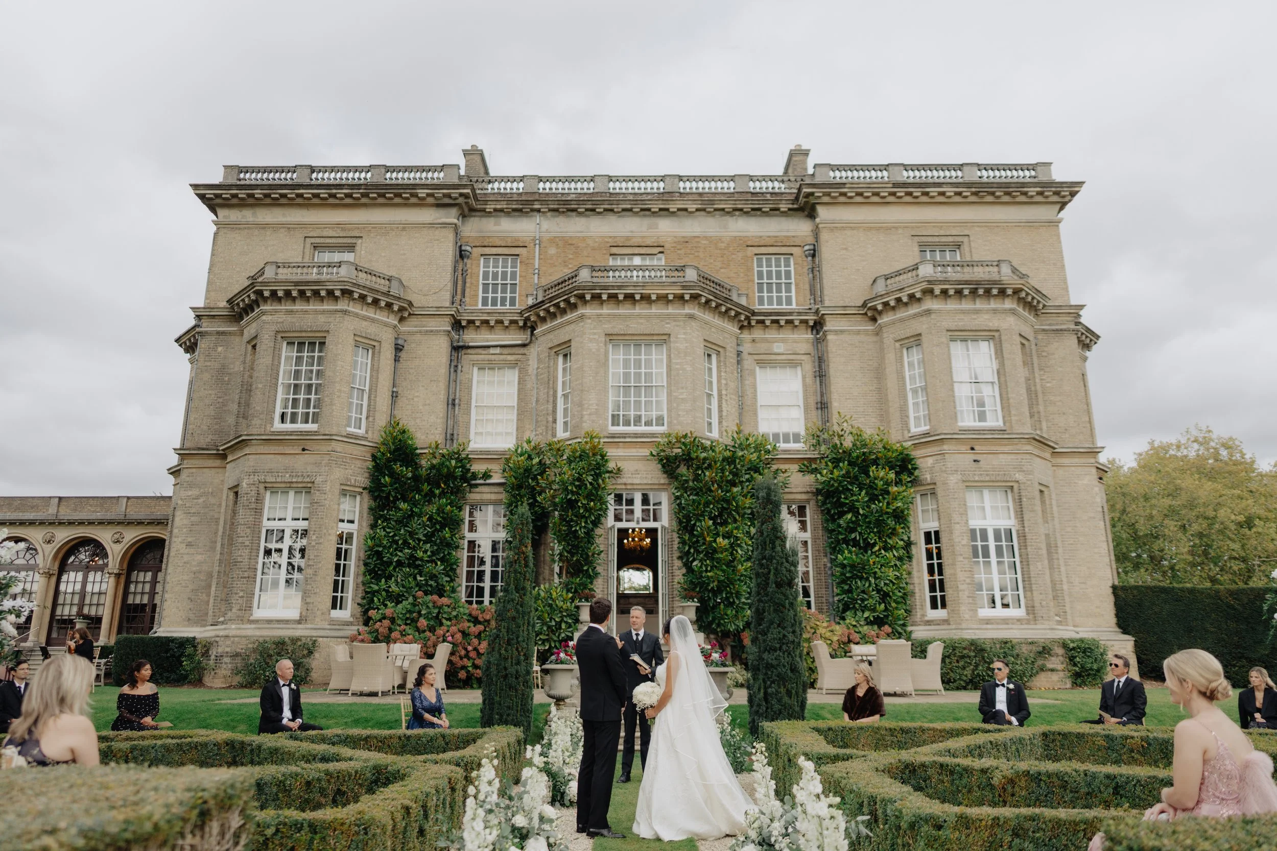 A wedding ceremony taking place outdoors in front of a large historic building with tall windows and ornate architecture. The bride and groom are standing in front of an officiant, surrounded by guests seated on the lawn. There are flowers and greenery decorating the garden area.