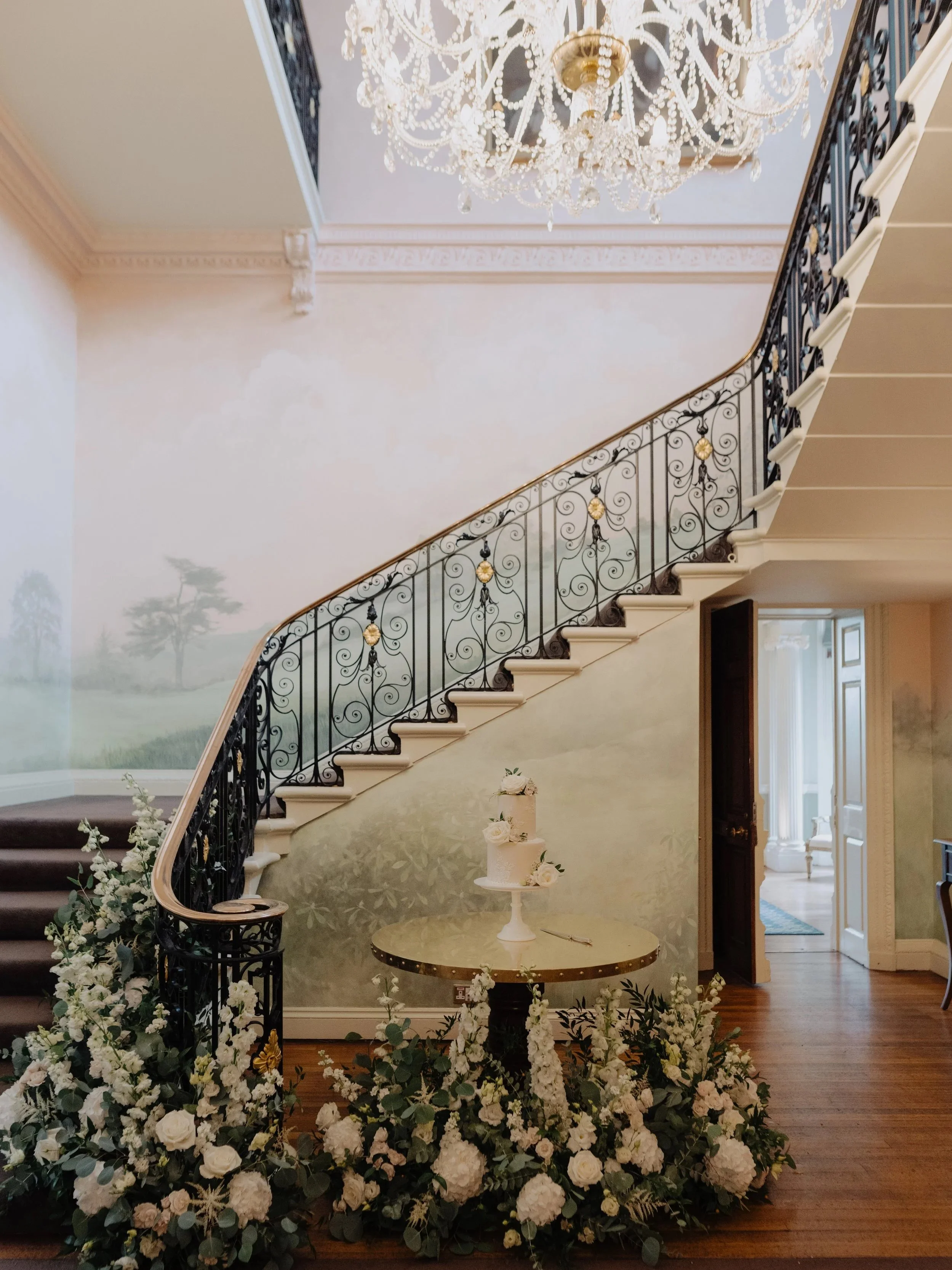 An elegant interior scene featuring a staircase with ornate black iron railing with gold accents, ornate crown molding, a large chandelier hanging from the ceiling, a table with a wedding cake decorated with flowers, and floral arrangements on the fl