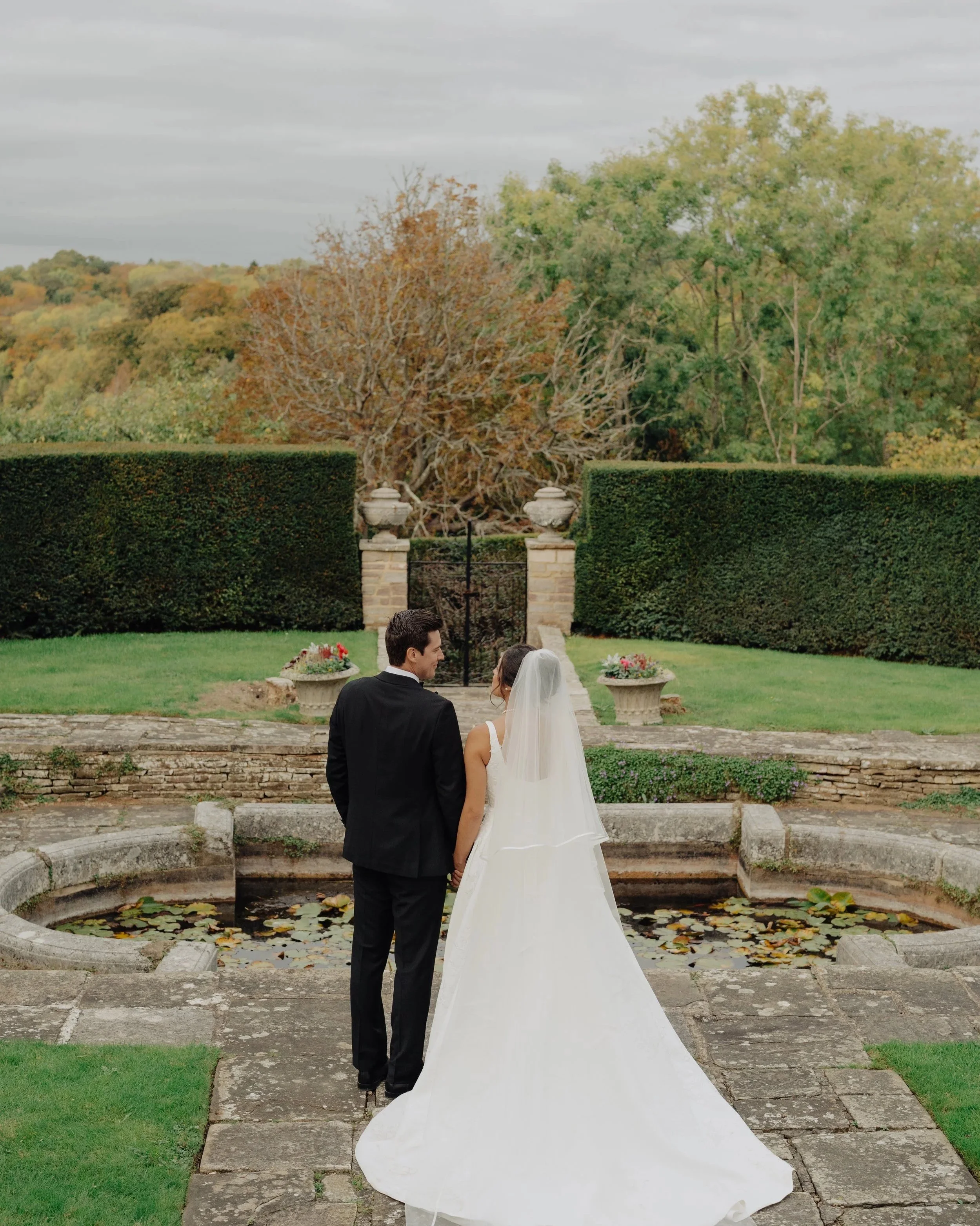 A bride and groom are holding hands and facing each other outdoors during a wedding ceremony. They are in front of a fountain with lily pads, surrounded by a stone pathway and greenery, with trees and hills in the background.