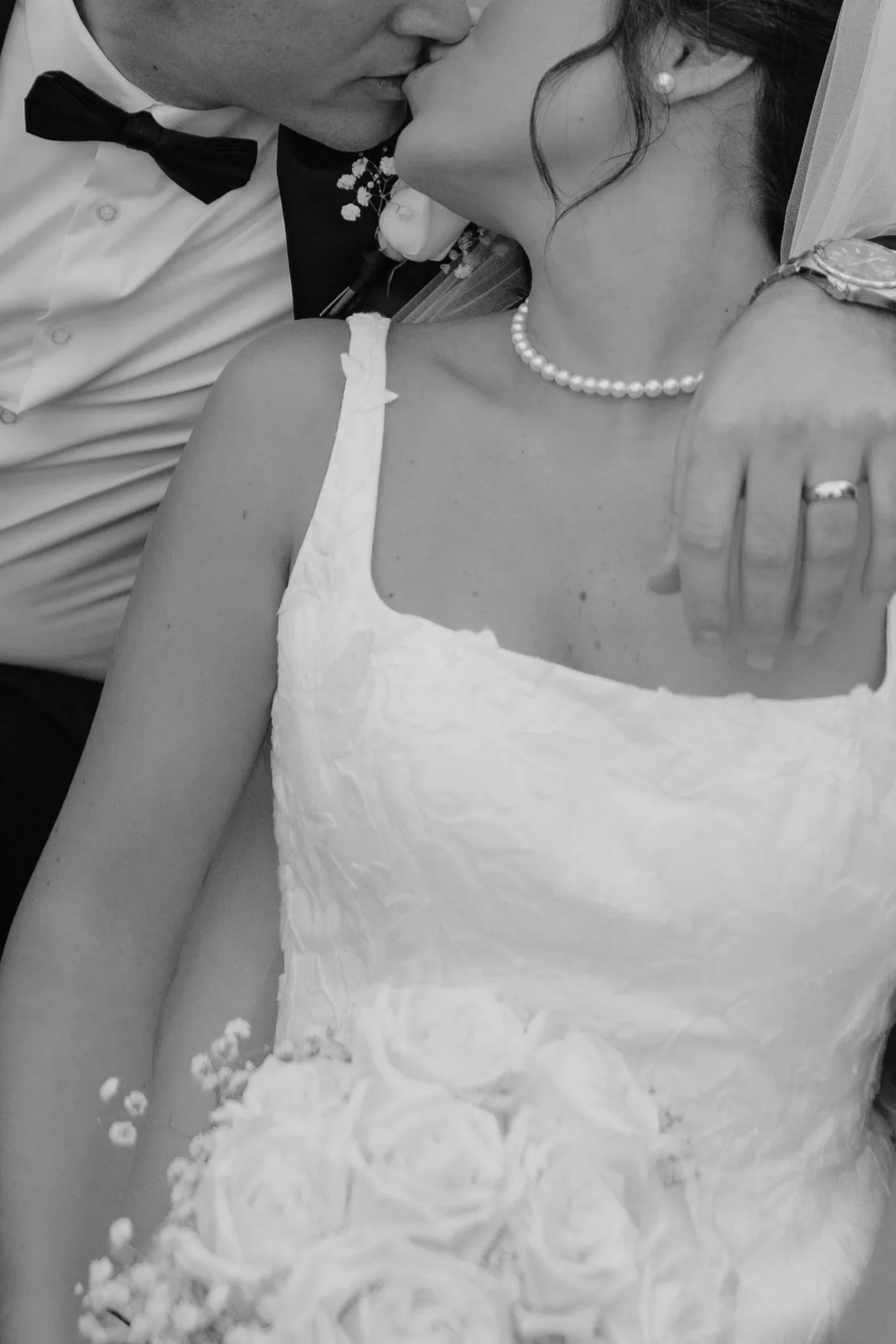 A black and white photo of a man and woman sharing a kiss. The woman is wearing a pearl necklace, earrings, and has a bracelet on her wrist. The man is dressed in formal attire with a bow tie. They are touching foreheads and lips.